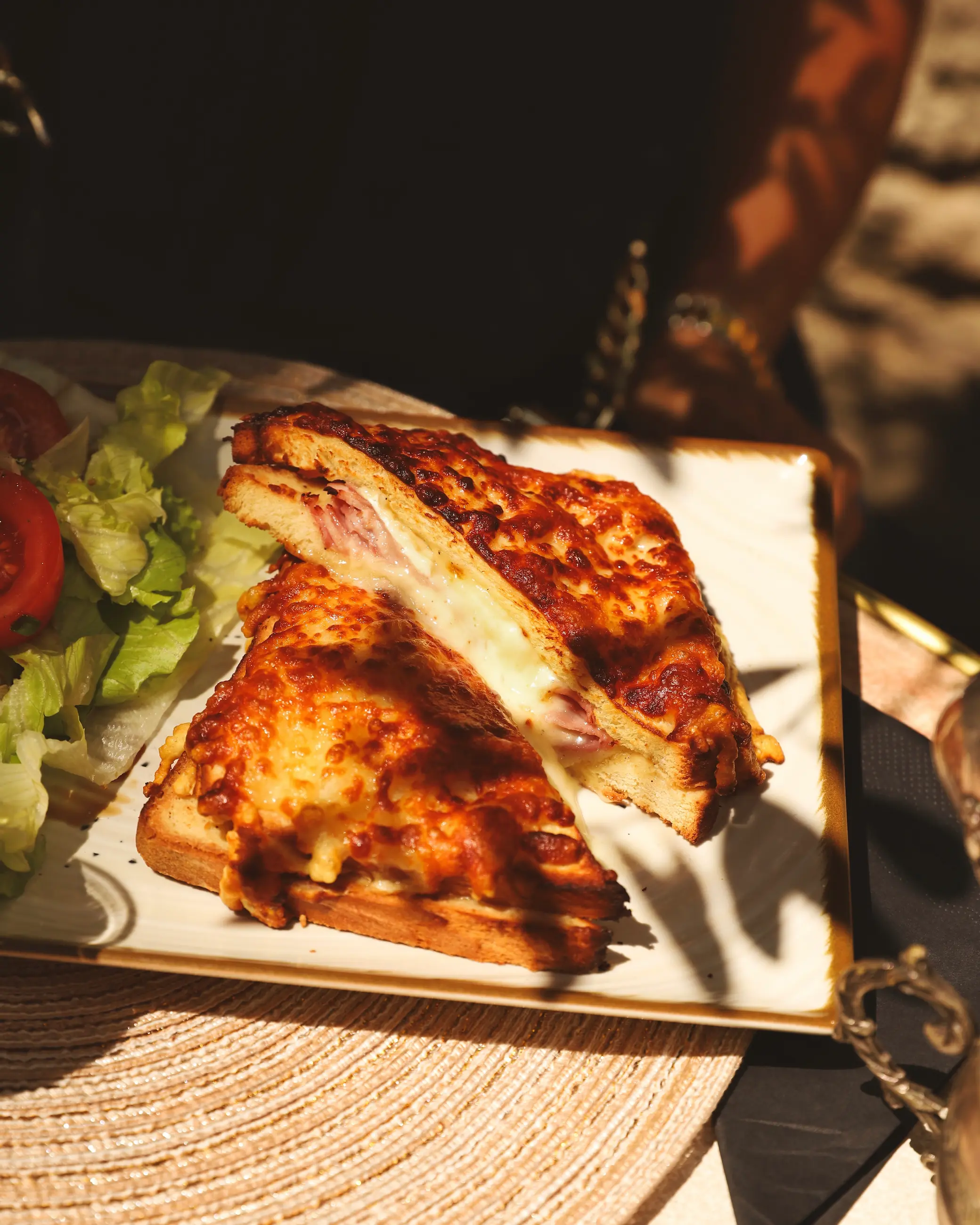 A golden, cheese-crusted croque monsieur sliced in half, oozing melted cheese and ham, served with a side salad of lettuce and tomato on a sunlit plate