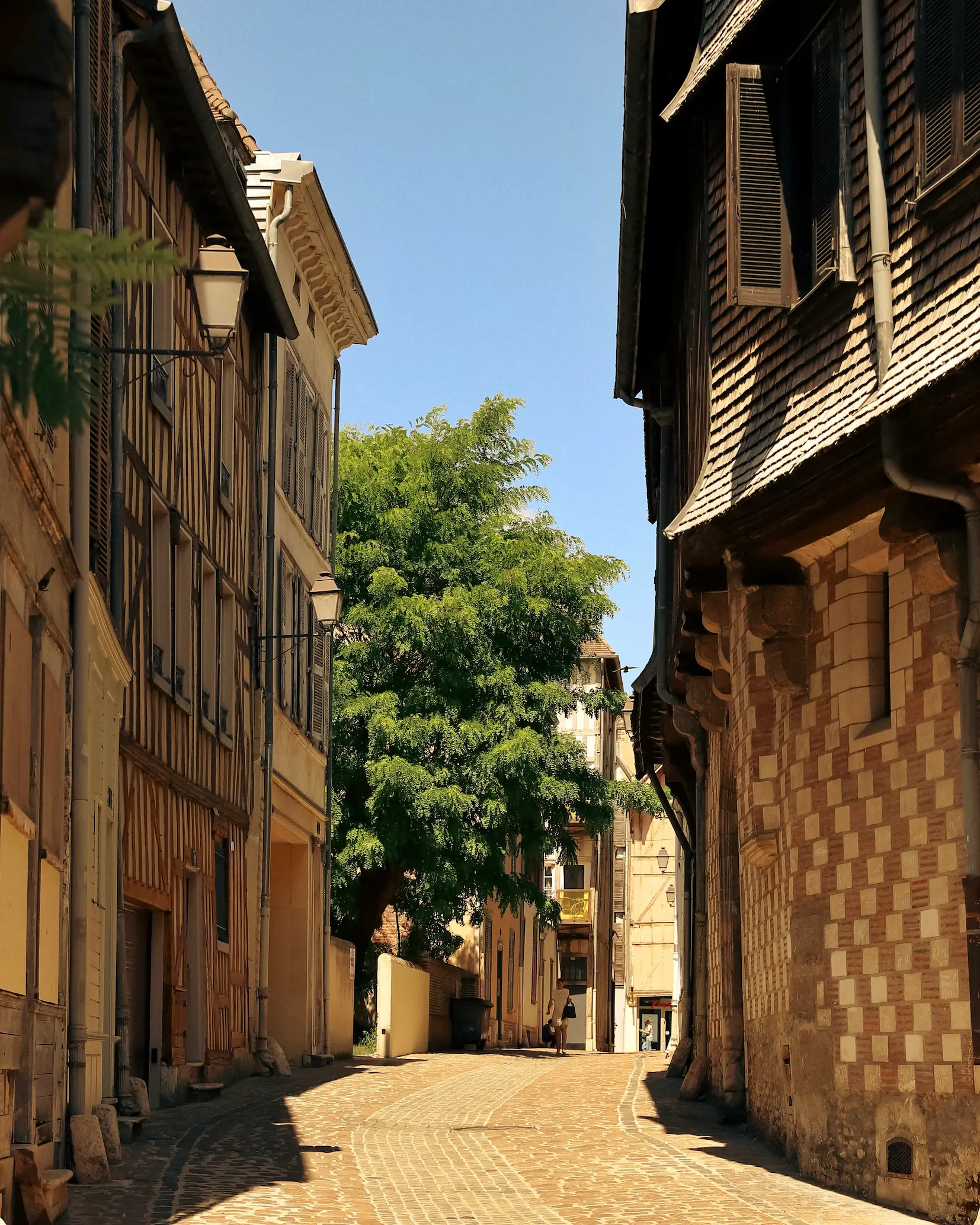 A sunlit cobbled street in the old town of Troyes, France, lined with medieval half-timbered houses and historic stone buildings, capturing the town's charming medieval architecture and peaceful atmosphere.