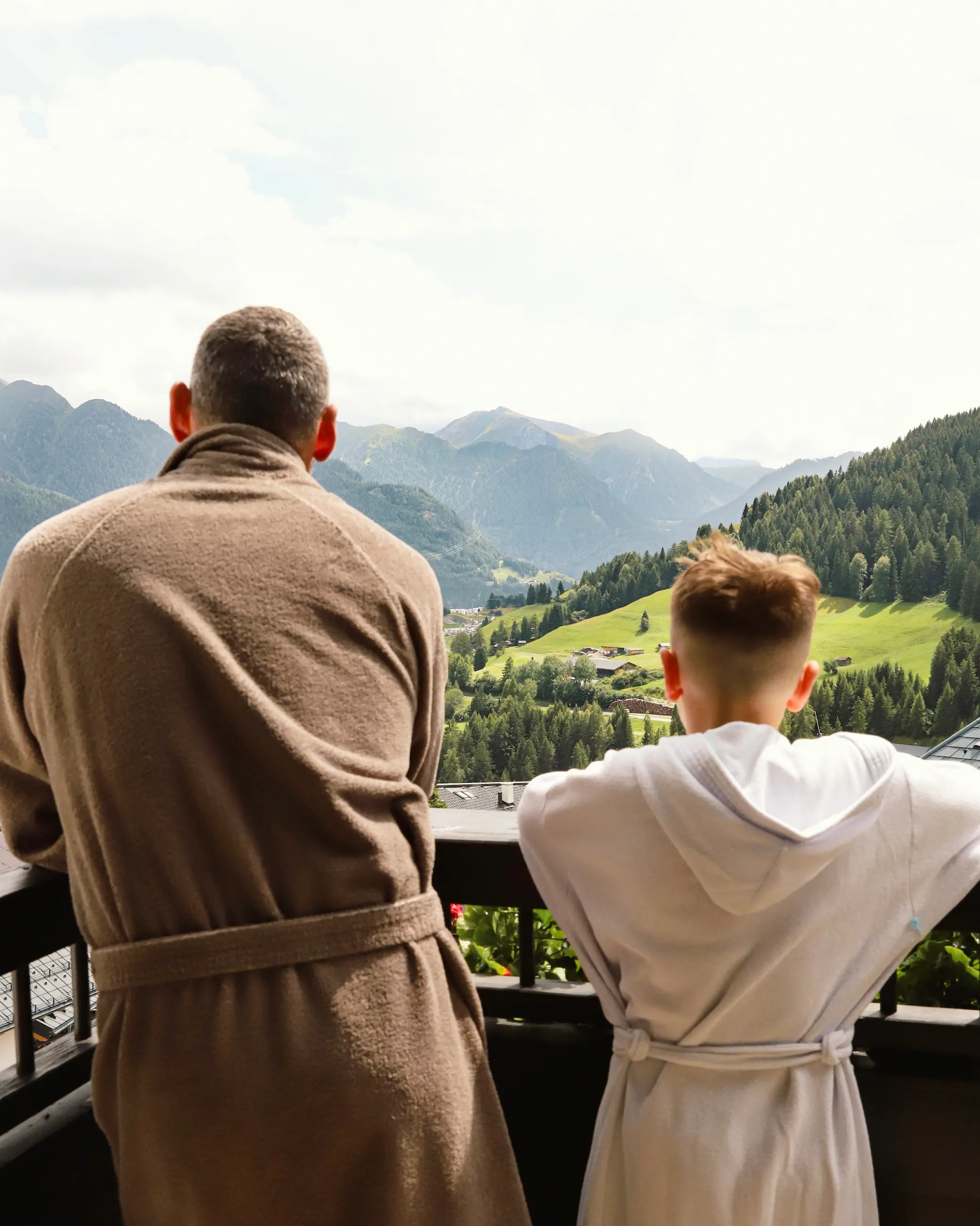 Two people in robes enjoy a panoramic balcony view of the lush green valley and mountain peaks in Fiss, Austria, from Schlosshotel Fiss.