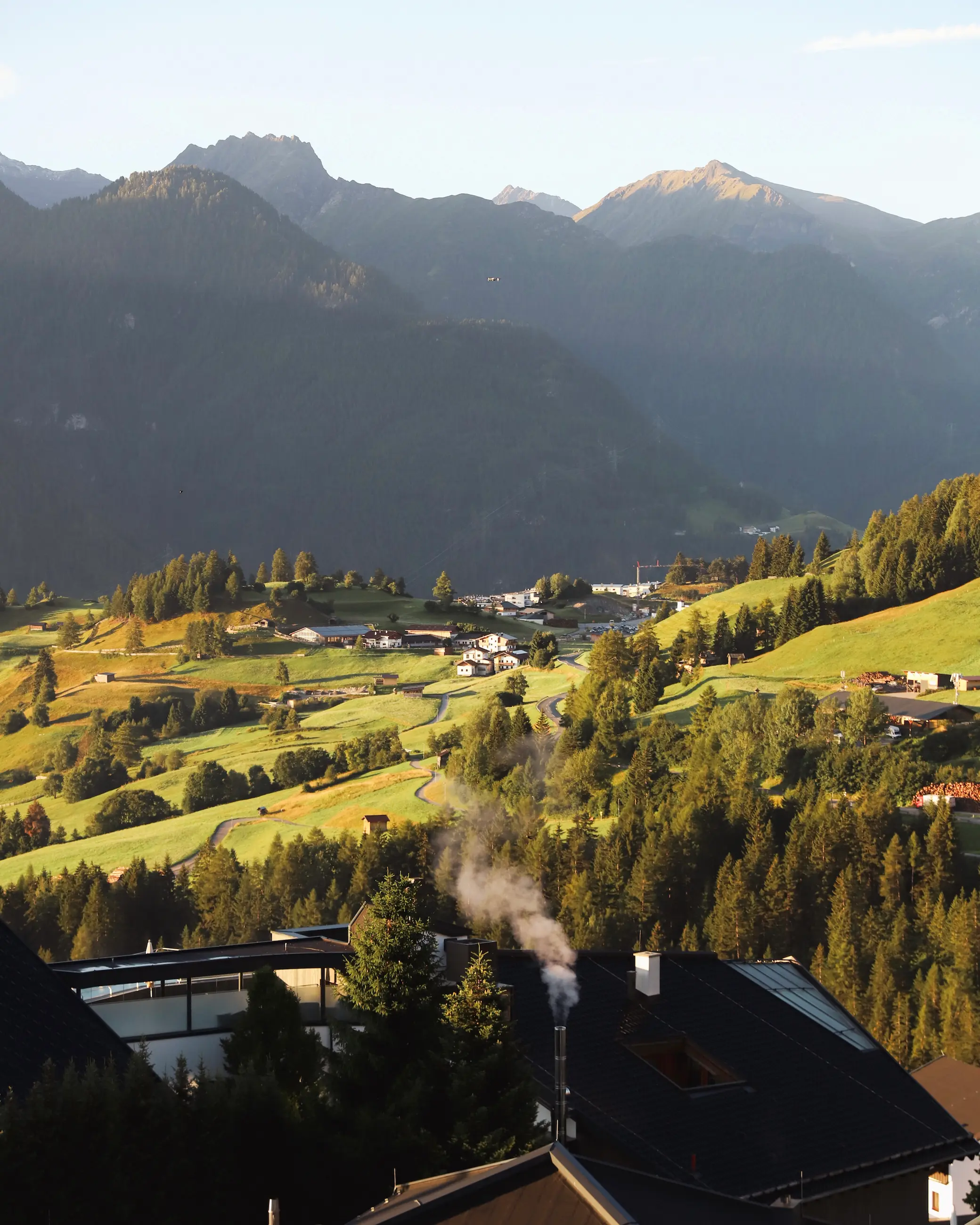 Morning sunlight over the green alpine valley and rolling hills of Fiss, Austria, viewed from Hotel Schlossfiss with a plume of chimney smoke rising into the crisp mountain air.