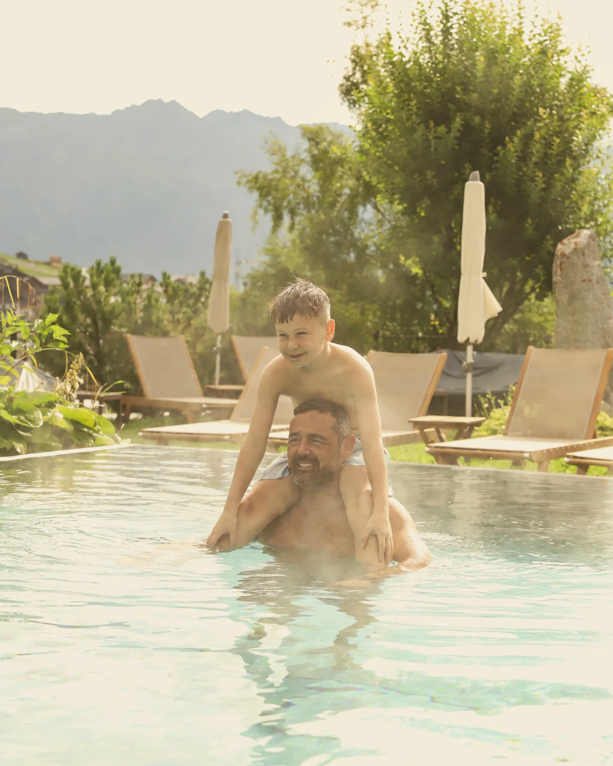 Father and son enjoying a playful moment in the warm outdoor pool at Schlosshotel Fiss, surrounded by loungers and mountain views in the Austrian Alps.