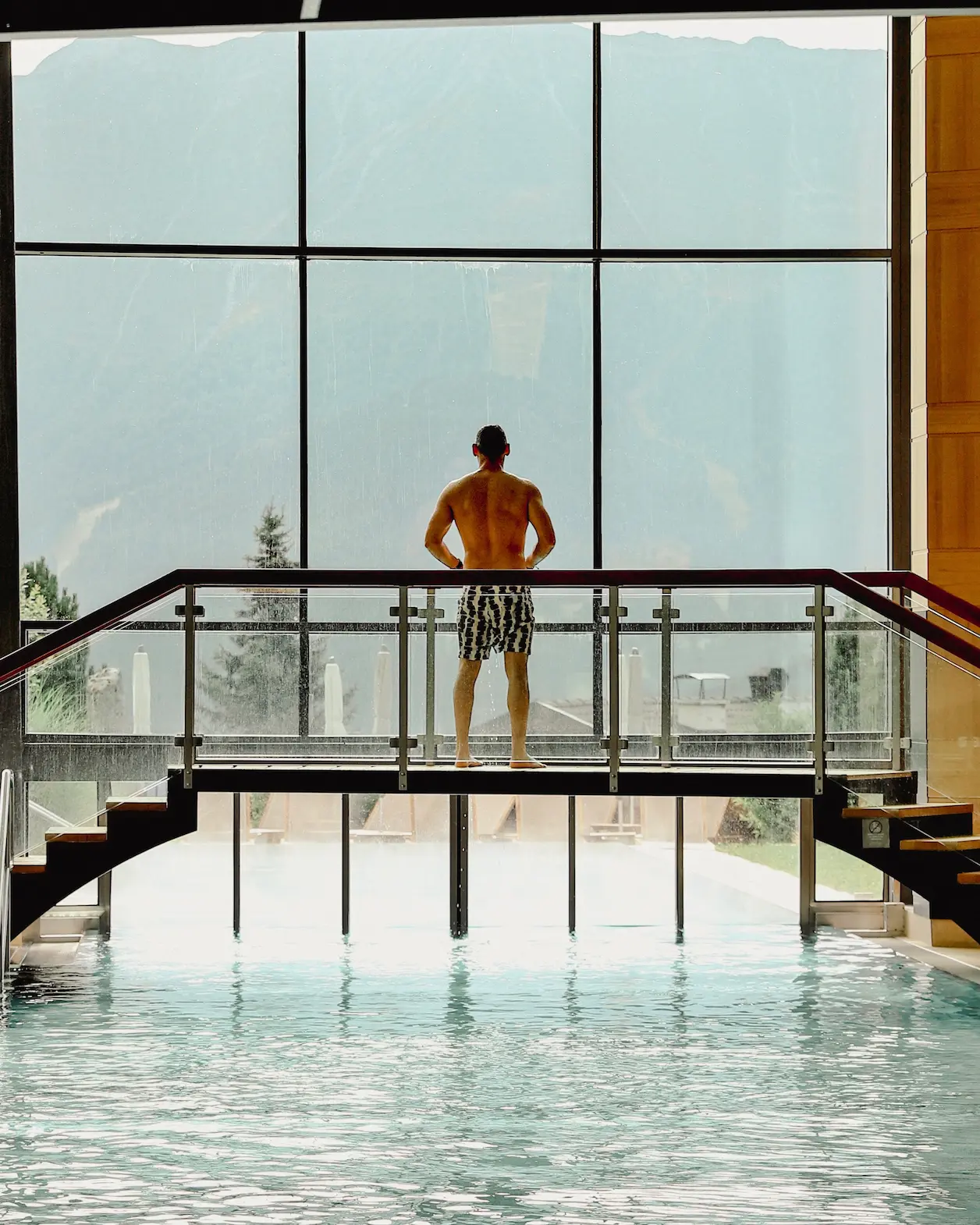 Man standing on a glass bridge overlooking the indoor-outdoor pool at Schlosshotel fiss, with a panoramic view of the Austrian Alps beyond