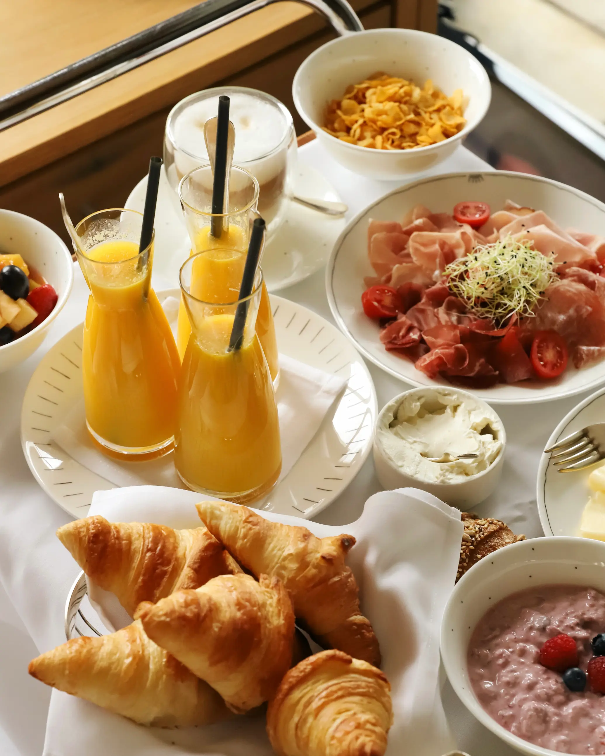 Continental breakfast spread at Schlosshotel Fiss featuring fresh croissants, fruit salad, cold cuts, soft cheeses, granola, vibrant orange juice, and cappuccinos — all served on a white tablecloth with mountain light streaming in.