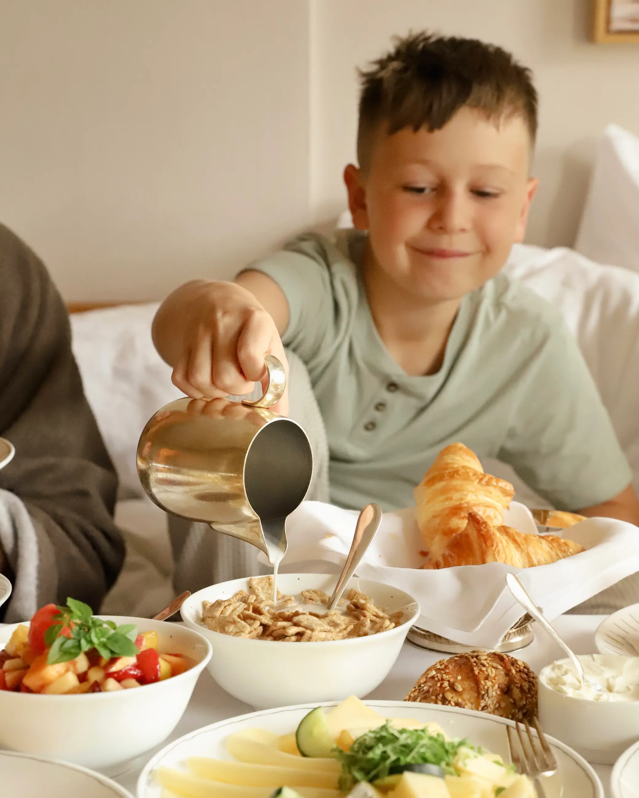 Young boy smiling while pouring milk onto cereal during a relaxed in-room breakfast at Schlosshotel Fiss, surrounded by fresh croissants, bread rolls, sliced cheese, fruit salad and yoghurt bowls – a cosy start to the day in the Austrian Alps.