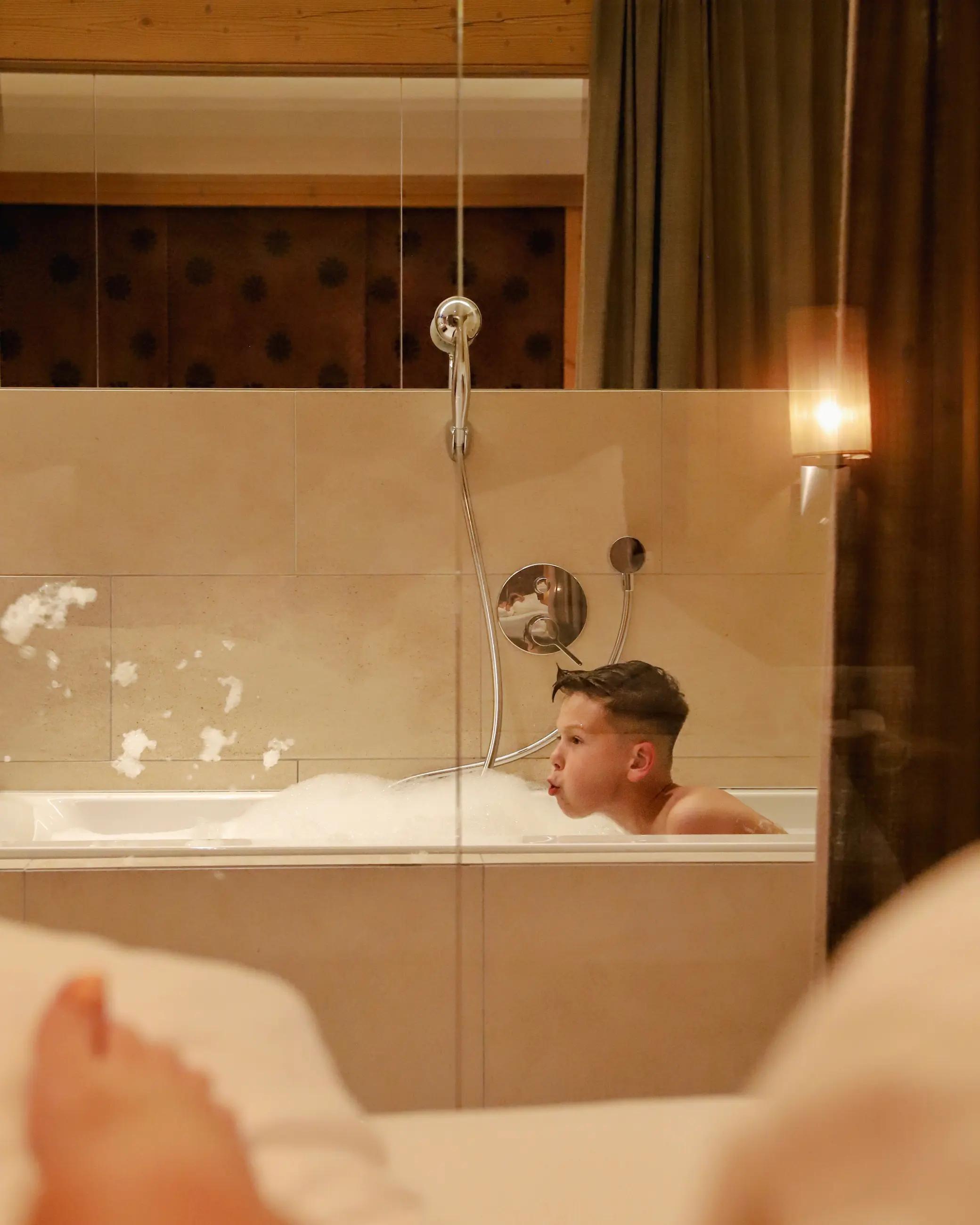 A child enjoying a bubble bath in a modern, cream-tiled bathroom with a floor-to-ceiling glass wall, seen from the lounge area of a family suite at Schlosshotel Fiss.