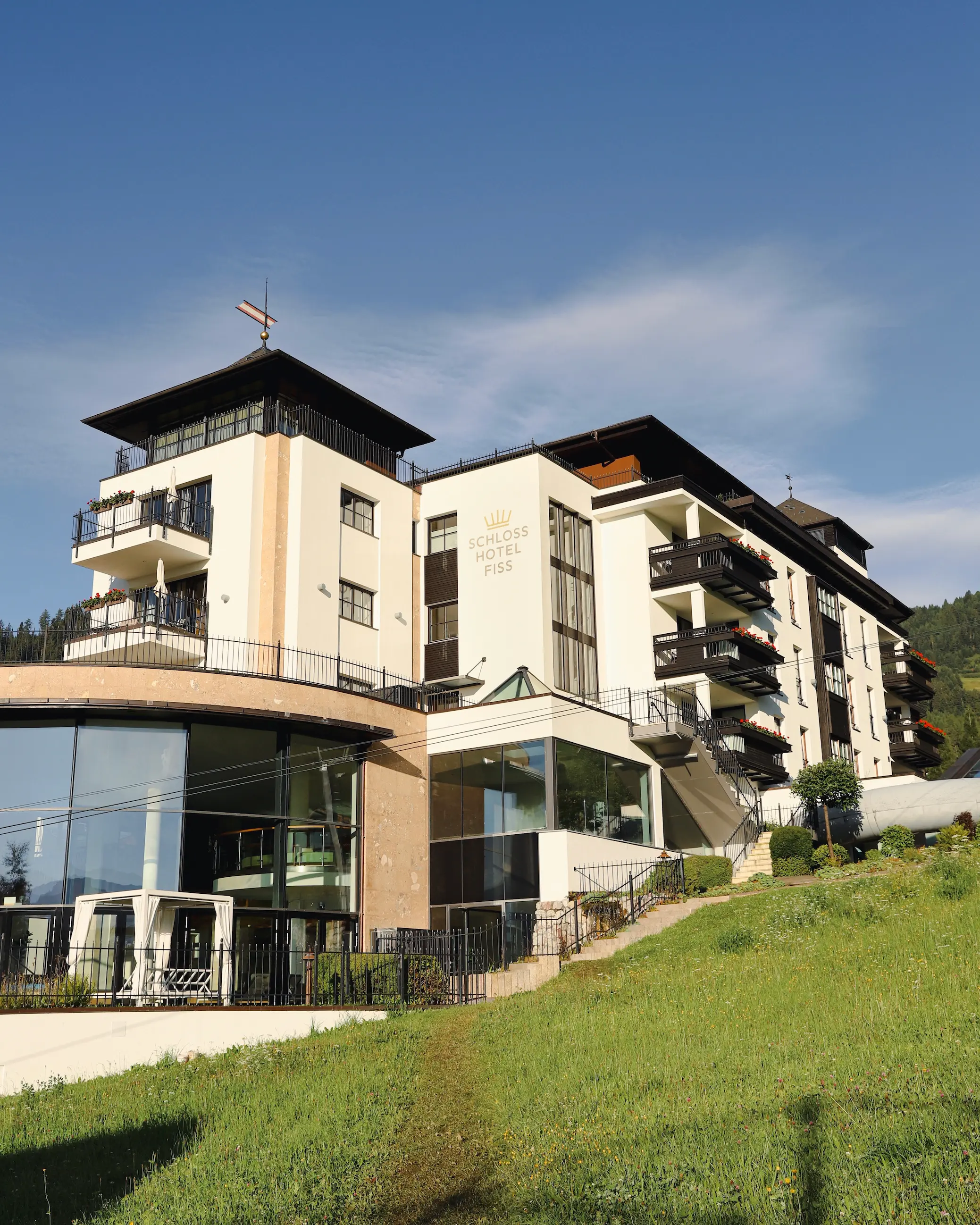 Exterior view of Schlosshotel Fiss in the Austrian Alps, featuring modern alpine architecture, balconies with flower boxes, and panoramic glass spa windows overlooking a grassy hillside.