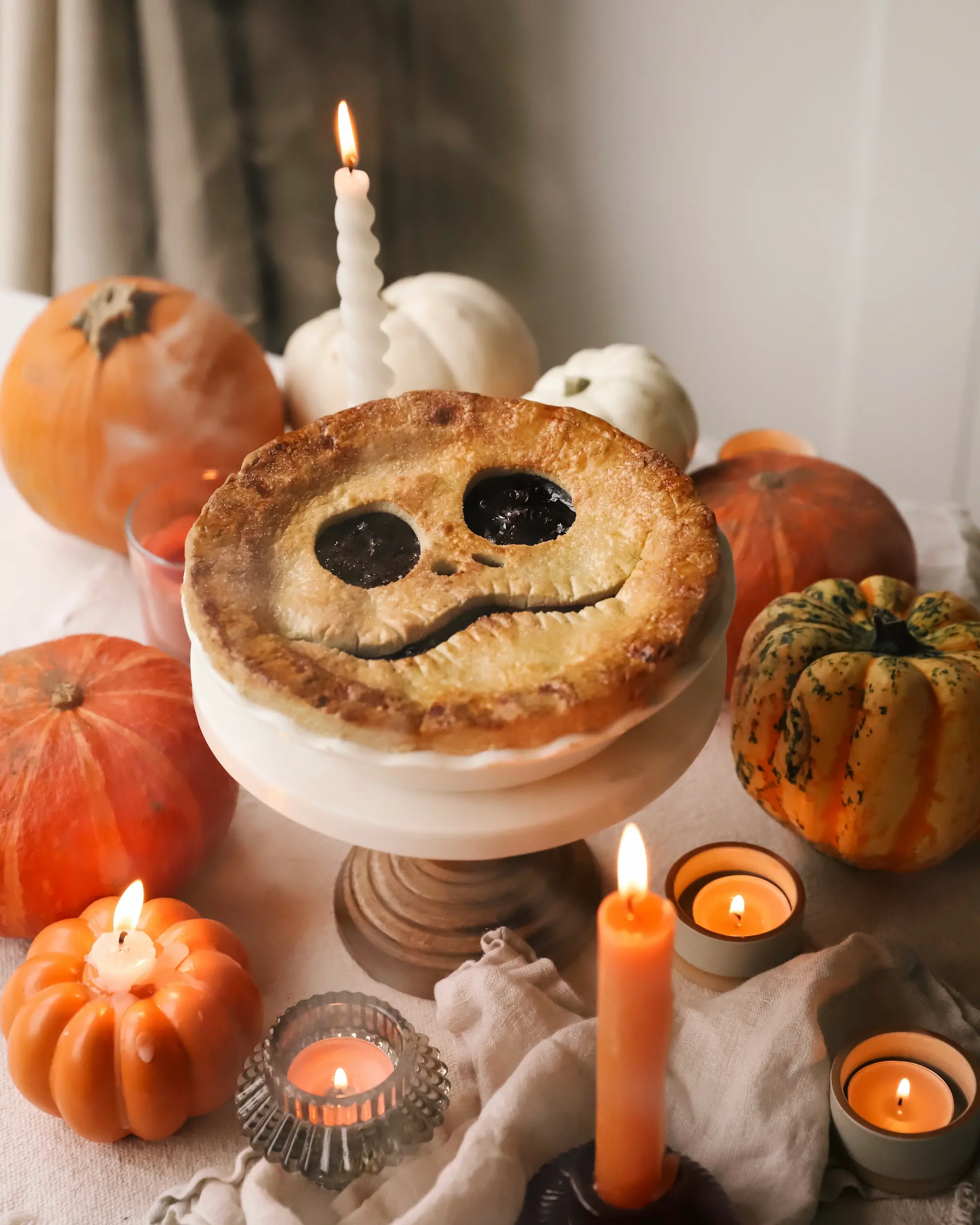 A golden, flaky cherry pie with Jack Skellington’s face cut into the crust, surrounded by glowing candles and small pumpkins on a warm, autumnal table setting.