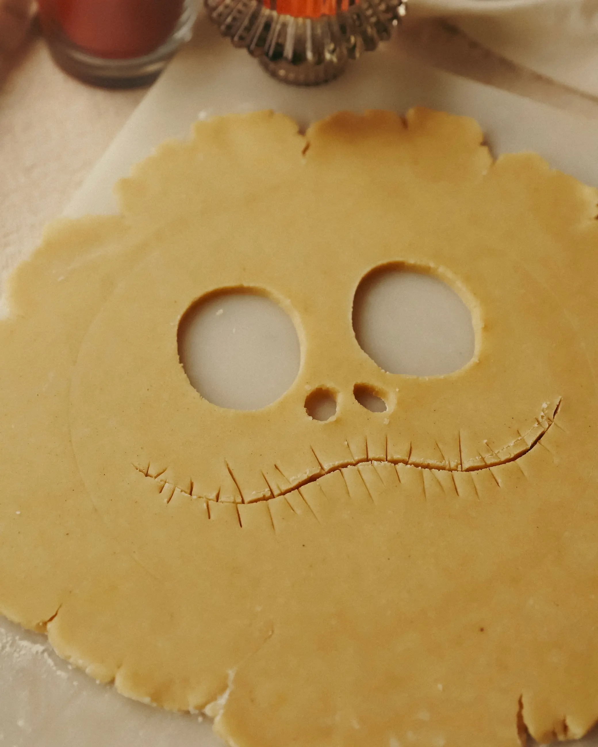 Close-up of rolled pie pastry with Jack Skellington’s face cut out — two round eyes, a small nose, and his stitched grin scored into the dough, ready for baking on a marble surface with warm candlelight nearby.