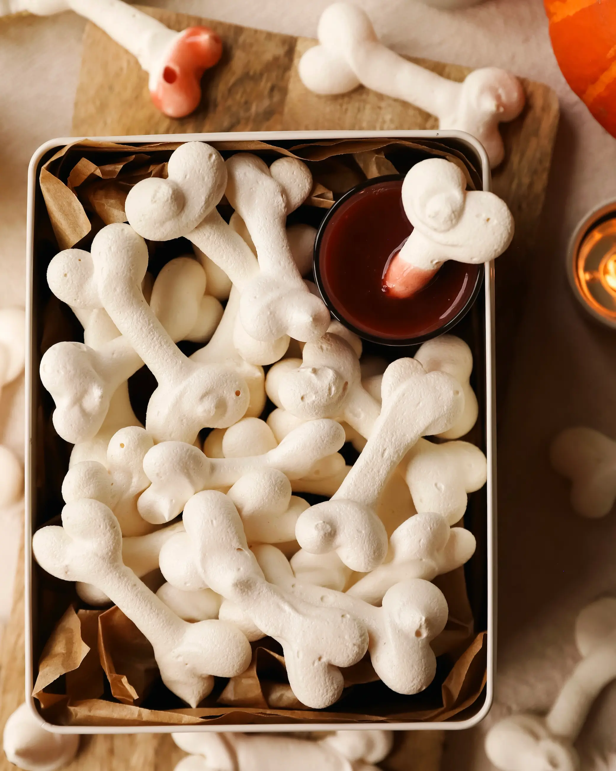 A Halloween-themed tin filled with crisp white meringue bones, served with a small pot of raspberry coulis “blood” for dipping, surrounded by candles and rustic props.