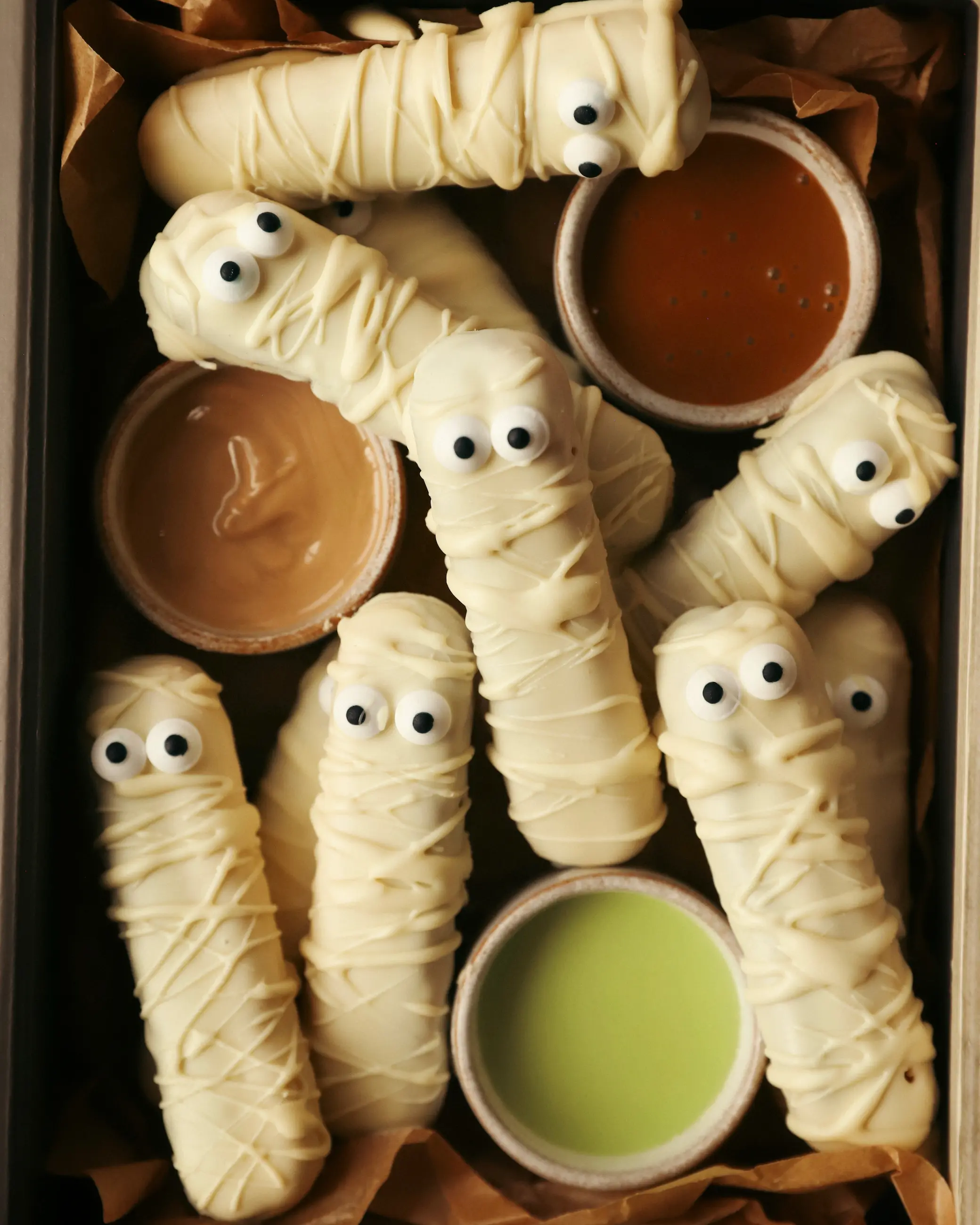 Close-up of white chocolate mummy dippers with candy eyes, arranged in a tray lined with parchment paper alongside bowls of caramel, pistachio, and hazelnut dipping sauces for a Halloween party spread.