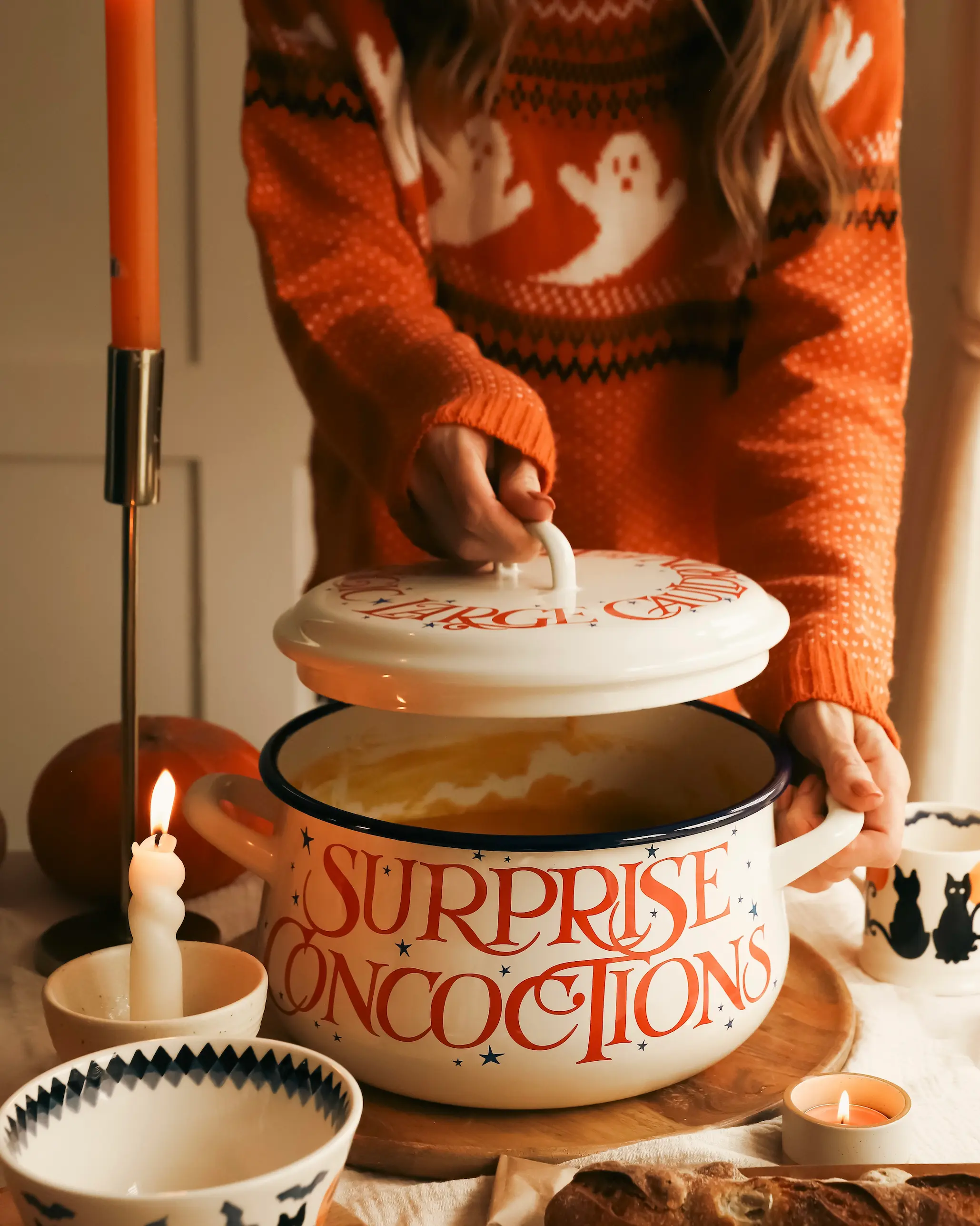 A person in an orange ghost-print Halloween jumper lifting the lid of a large cream enamel pot labelled “Surprise Concoctions,” filled with pumpkin soup. Surrounded by candles, pumpkins, and Halloween-themed bowls on a cosy autumn table.