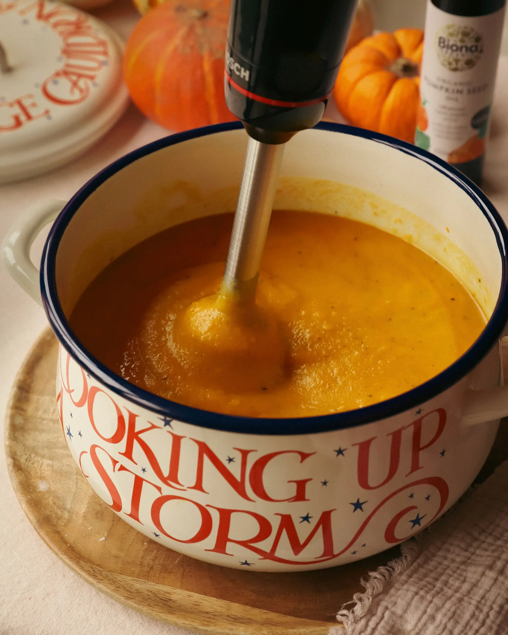 A hand blender mixing creamy roasted pumpkin and butterbean soup in a cream Emma Bridgewater enamel pot with red lettering that reads “Cooking Up a Storm,” surrounded by small pumpkins and a bottle of pumpkin seed oil on the table.
