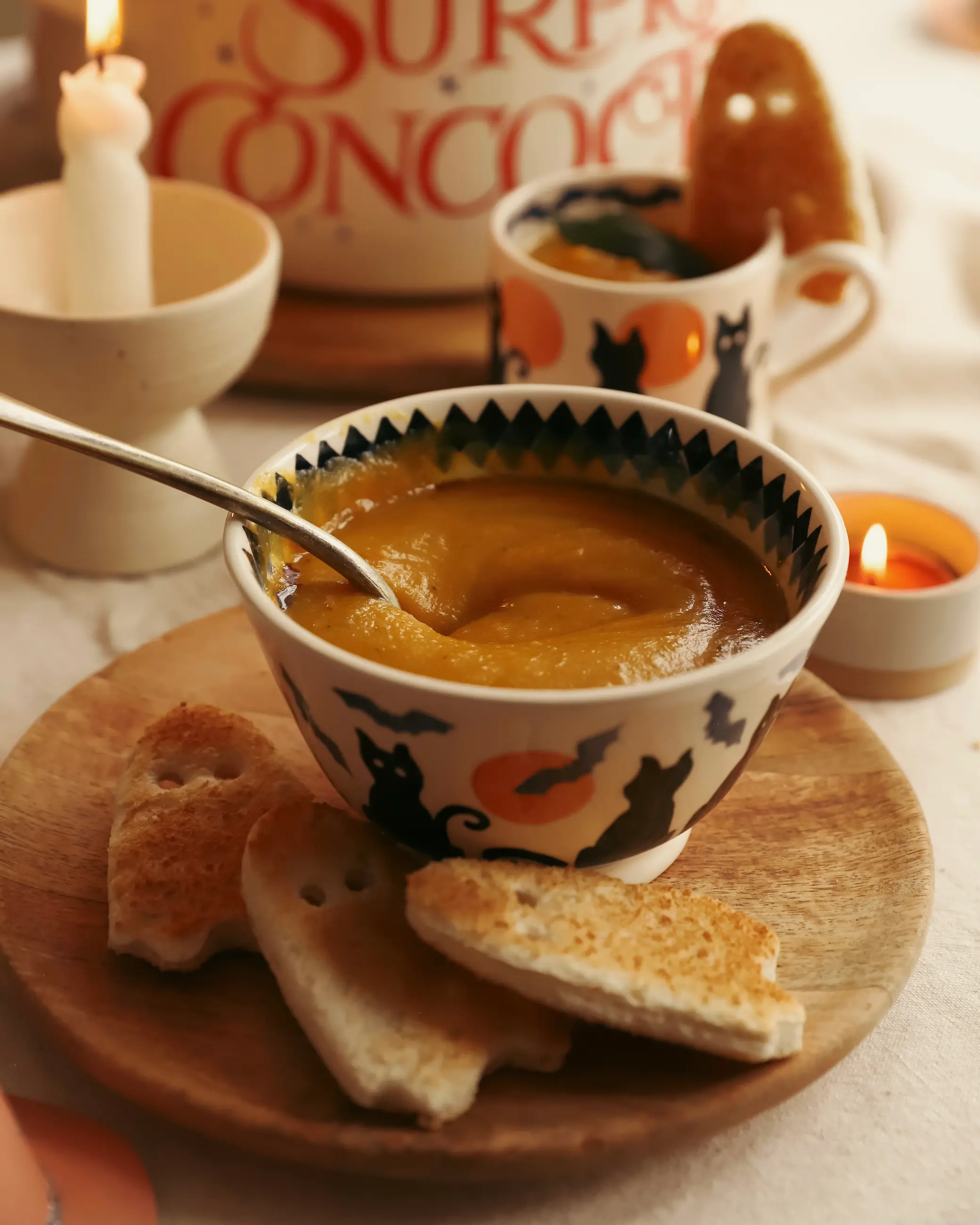 A bowl of creamy roasted pumpkin and butterbean soup served in a Halloween-themed mug with black cat and bat designs, on a wooden plate with toasted ghost-shaped bread. A cosy autumn table setting with candles glowing in the background.