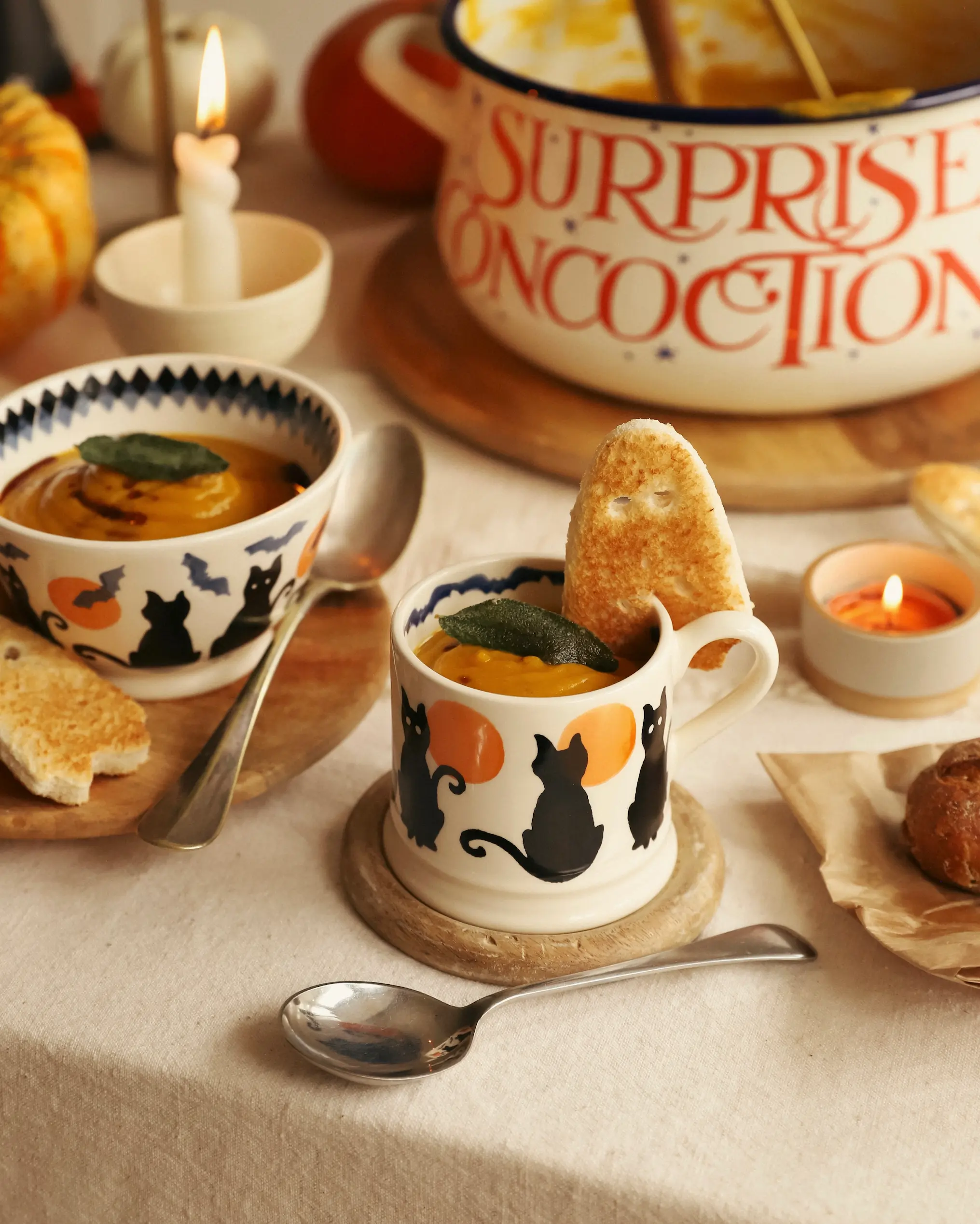 Emma Bridgewater Halloween mugs and bowls decorated with black cats and pumpkins, filled with creamy roasted pumpkin and butterbean soup. Served with ghost-shaped toast and surrounded by candles and autumnal pumpkins on a cosy table setting.