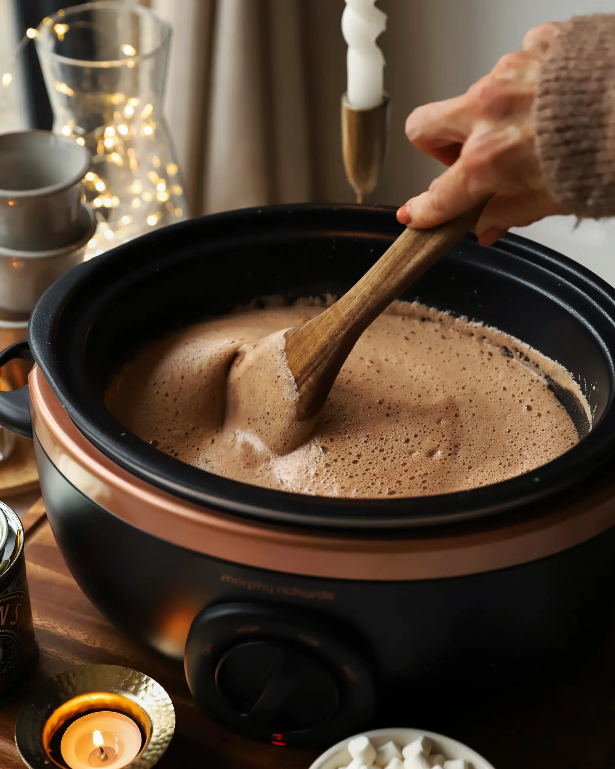 Person stirring creamy slow cooker hot chocolate with a wooden spoon, surrounded by mugs, mini marshmallows, and warm candlelight — a cosy winter drink scene with soft festive lighting.