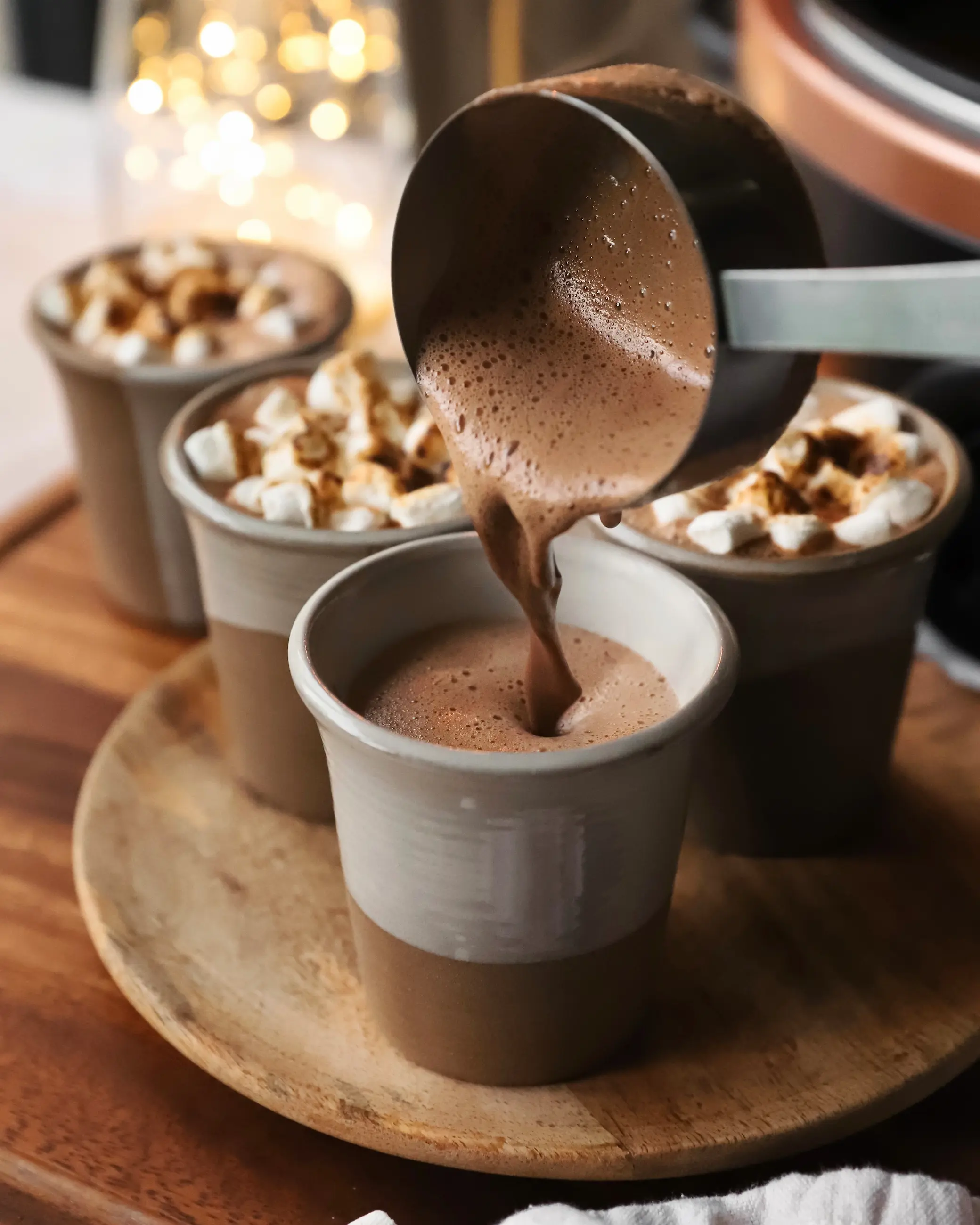 Pouring rich, frothy slow cooker hot chocolate into ceramic mugs, topped with toasted mini marshmallows and surrounded by soft festive lights.