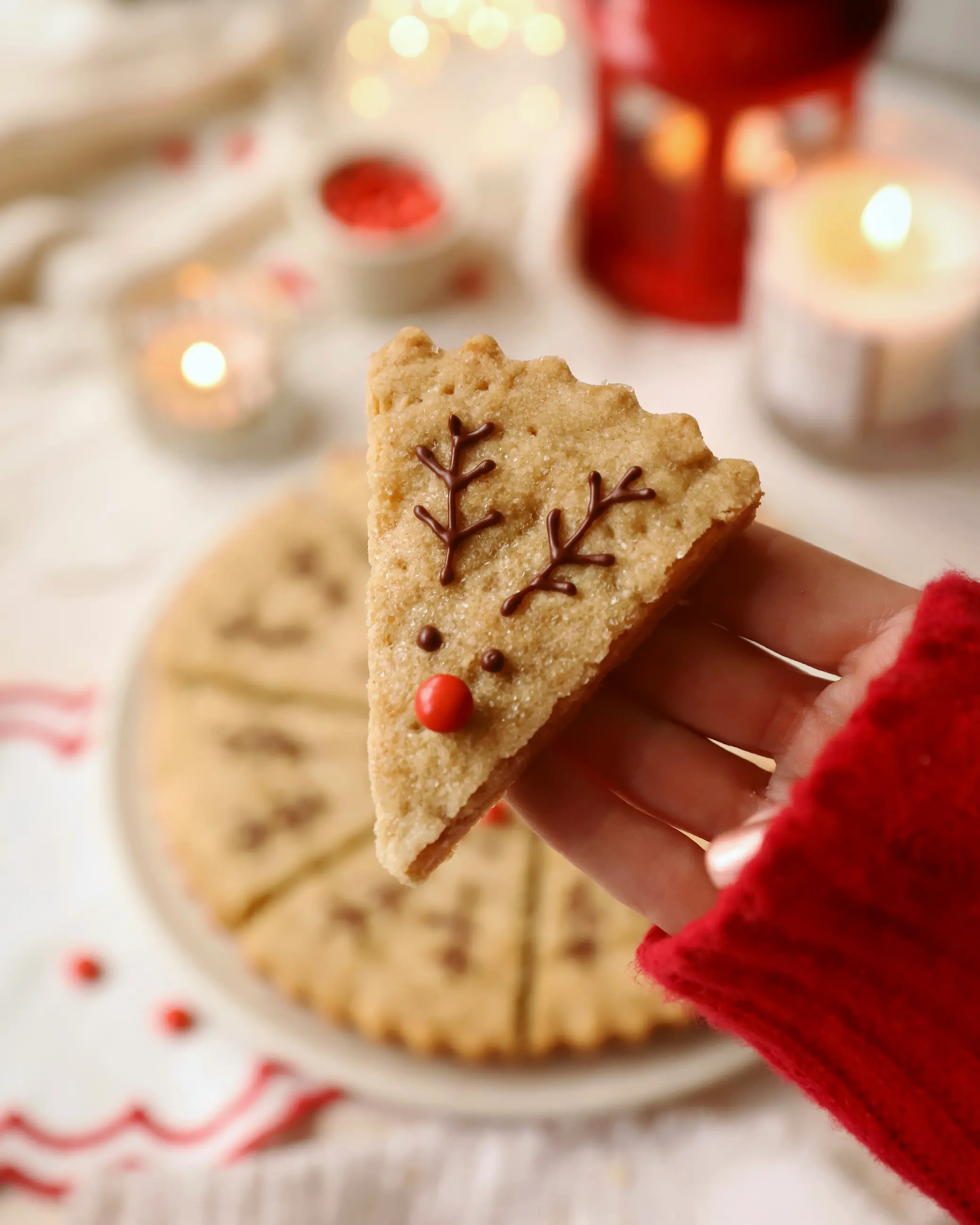 A hand holding a festive reindeer-shaped gingerbread maple shortbread wedge, decorated with chocolate antlers and a red nose. A full round of shortbread slices sits blurred in the background, surrounded by warm candles and soft Christmas lights for a cosy holiday feel.