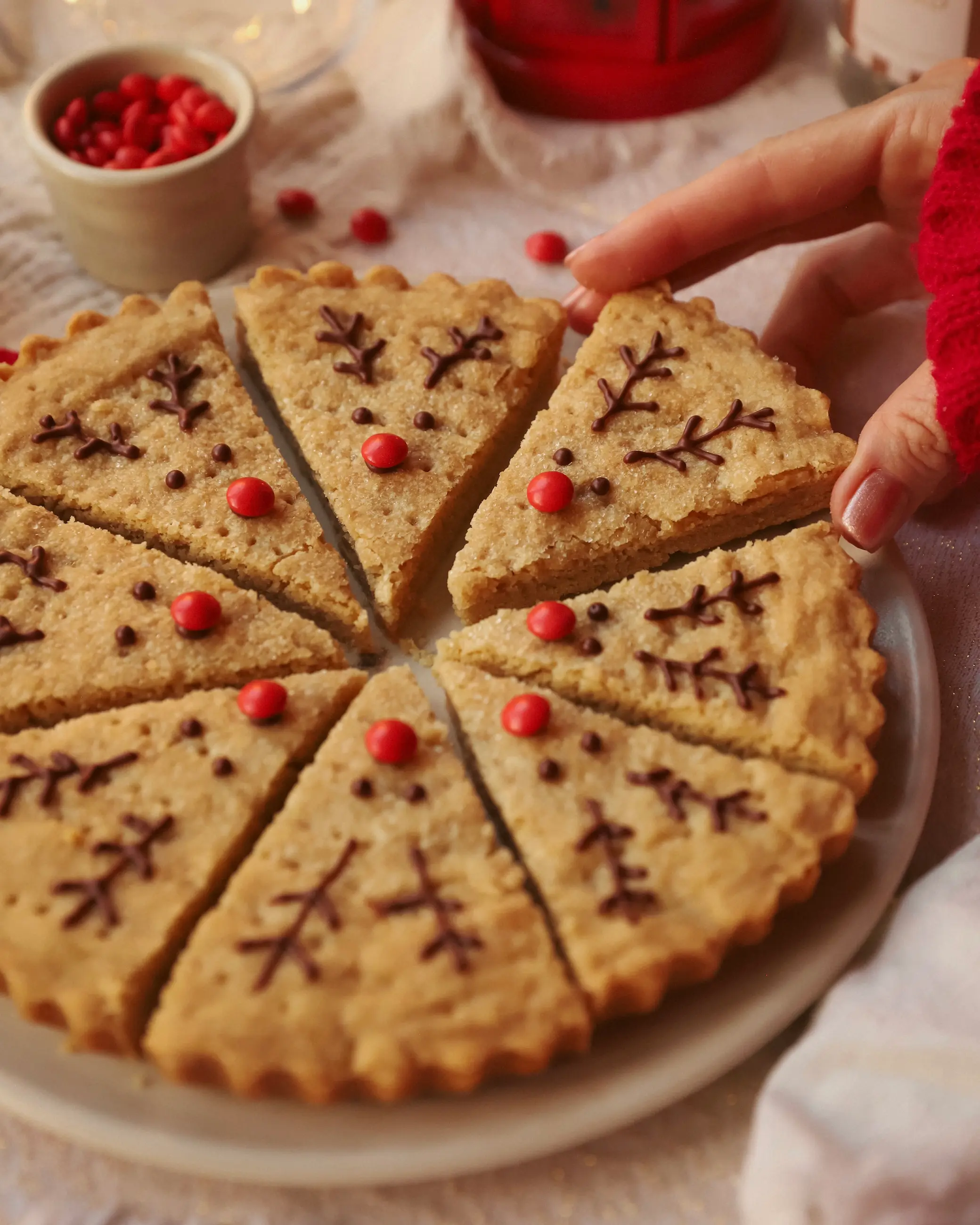 A hand in a red cosy sleeve lifting one reindeer-decorated gingerbread maple shortbread wedge from a full round of cut shortbread. Each slice features chocolate antlers and a red candy nose, with festive red sweets and warm candlelit tones in the background for a soft, Christmassy atmosphere.