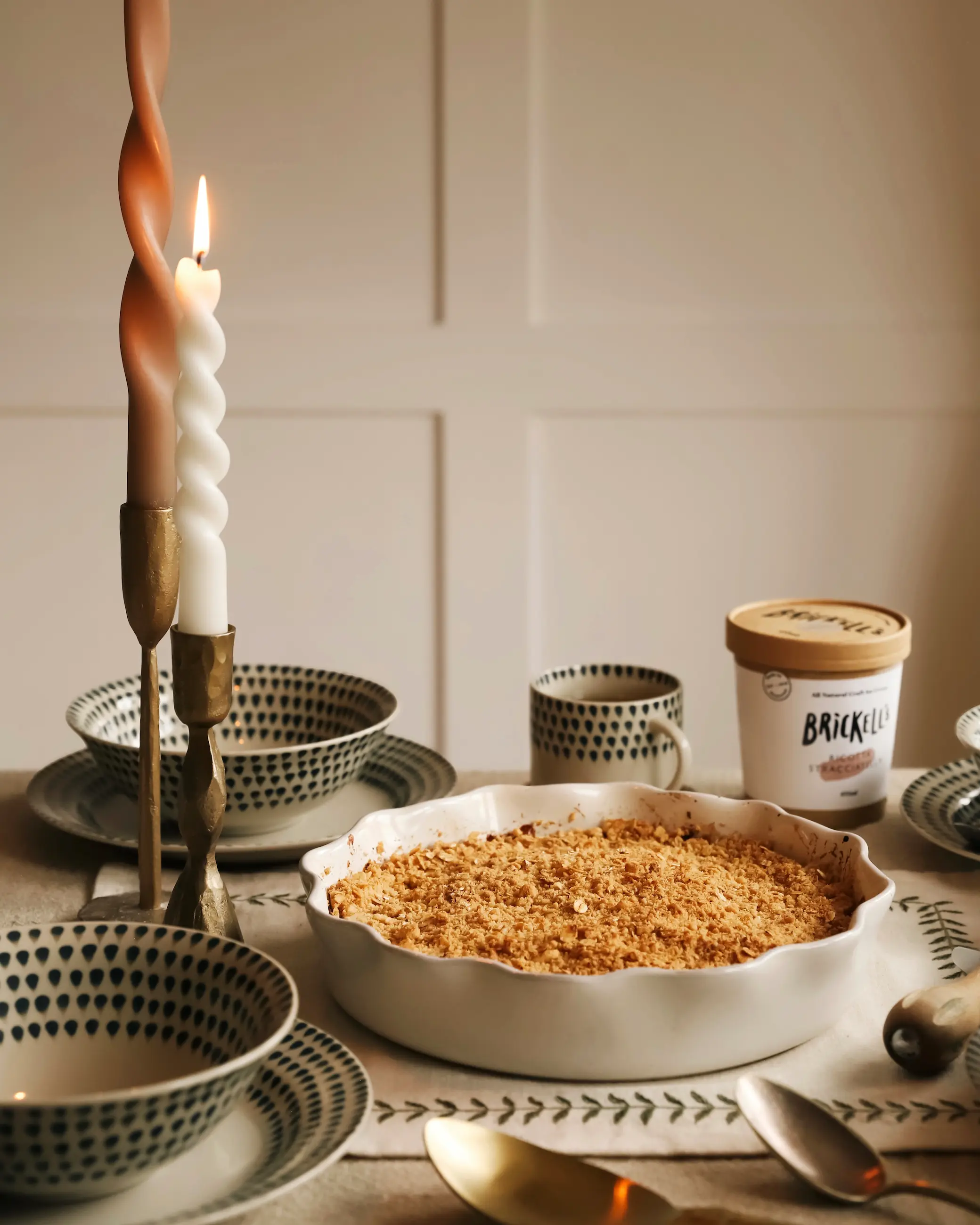 A baked apple crumble in a white ceramic dish sits on a dining table, surrounded by patterned bowls, spoons, a lit candle and a tub of ice cream in the background.