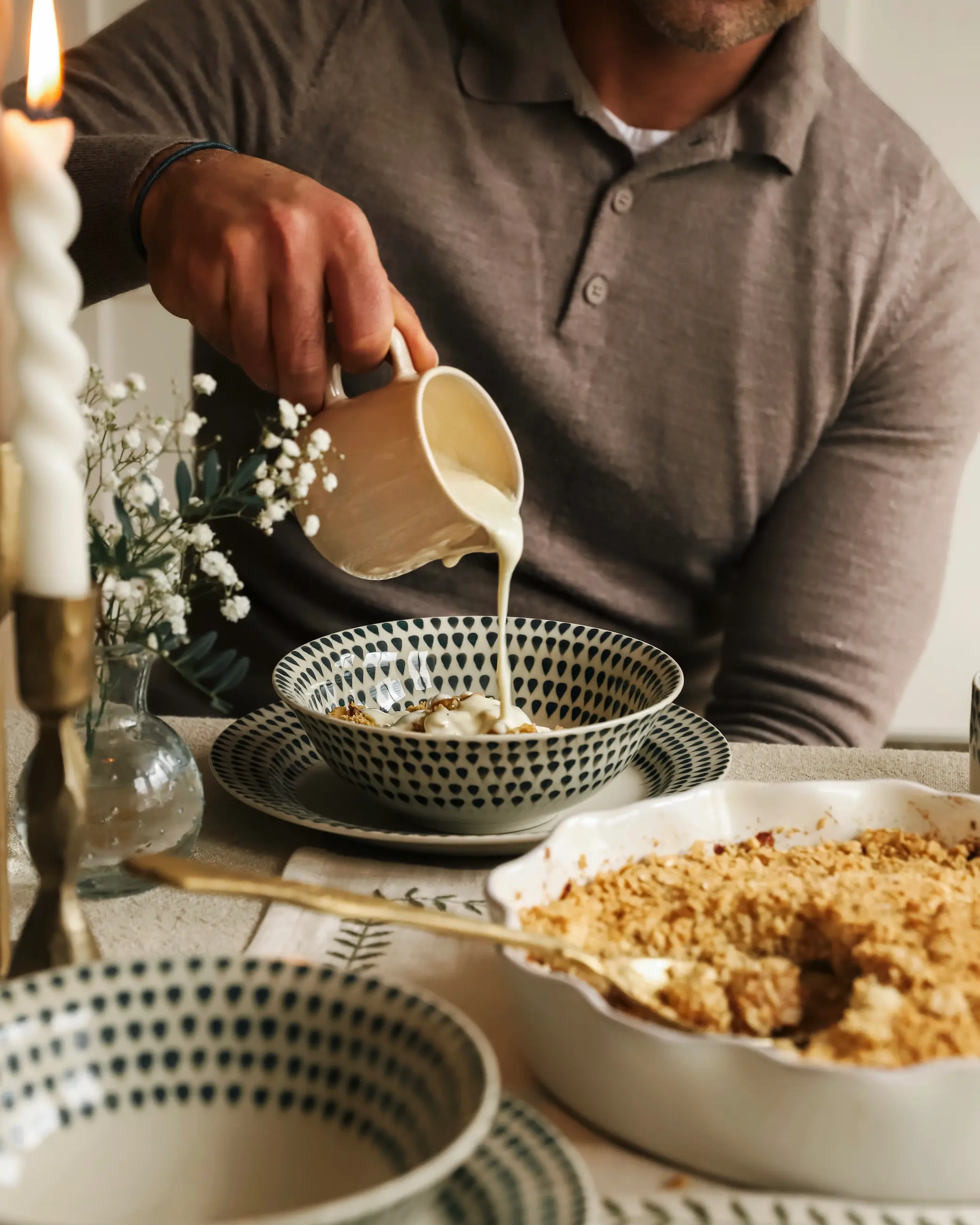 Cream is being poured from a small jug into a patterned bowl of apple crumble, with a baking dish of crumble on the table beside it and a lit candle in the background.