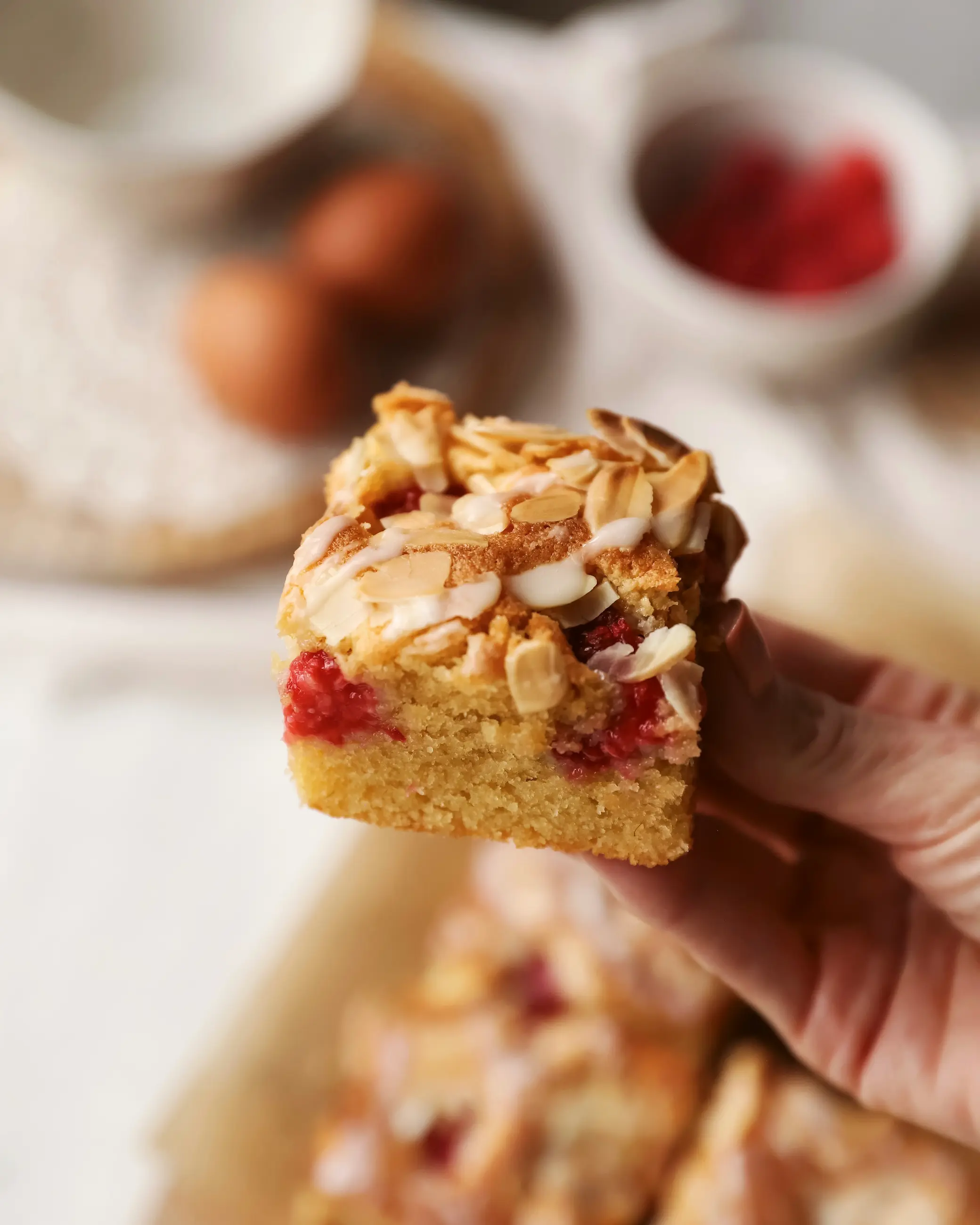 Hand holding a square of raspberry and almond traybake with fresh raspberries baked into a soft golden sponge, topped with flaked almonds and a light drizzle of icing, with the rest of the traybake blurred in the background.