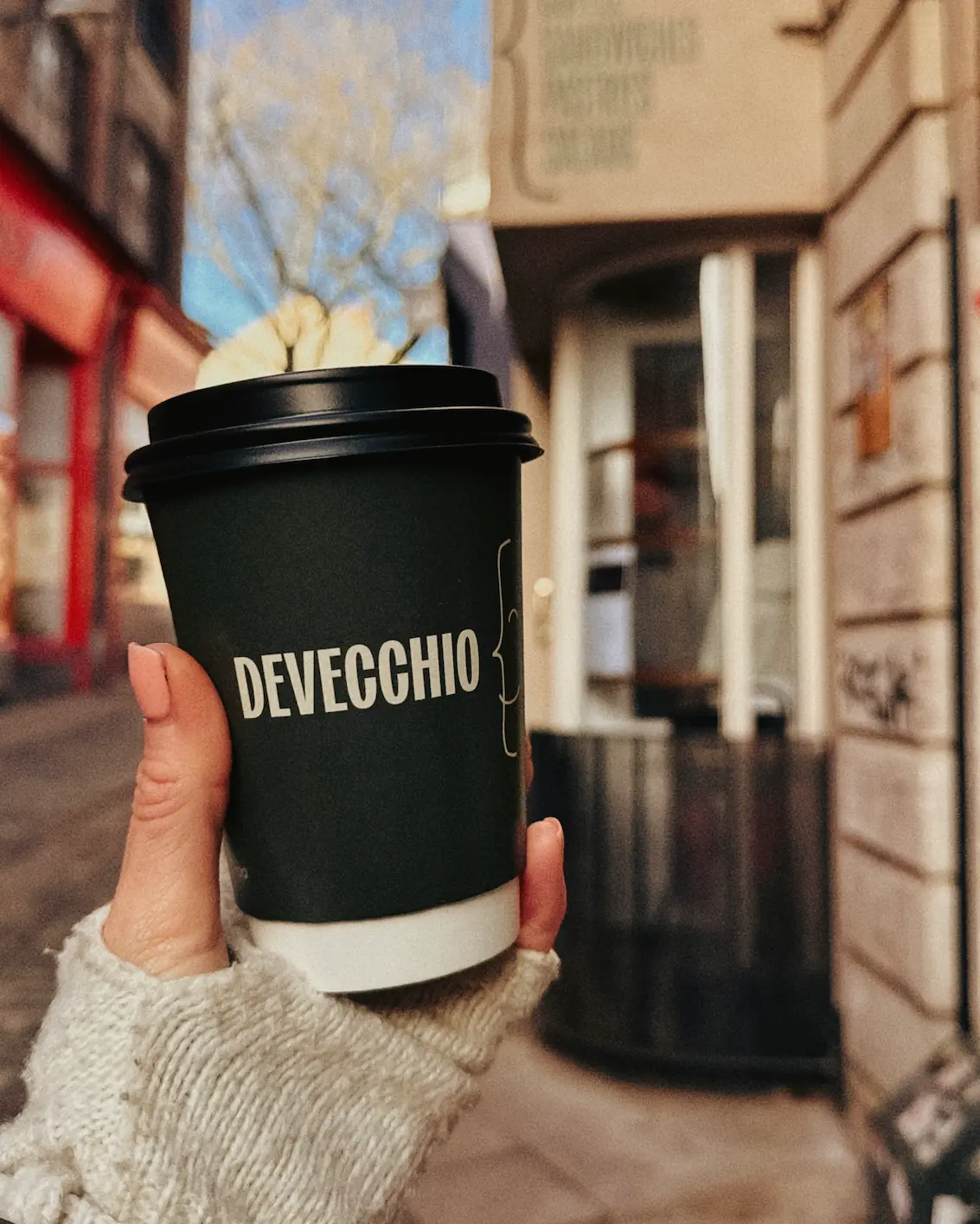 Hand holding a takeaway coffee from Devecchio bakery on a Norwich street, with blurred city buildings in the background.