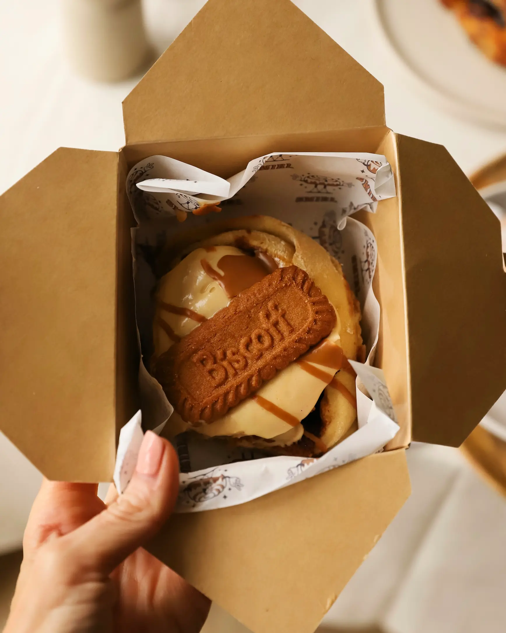 Hand holding a takeaway pastry box from Swirl bakery in Norwich, filled with a Biscoff-topped cinnamon bun drizzled with sauce.