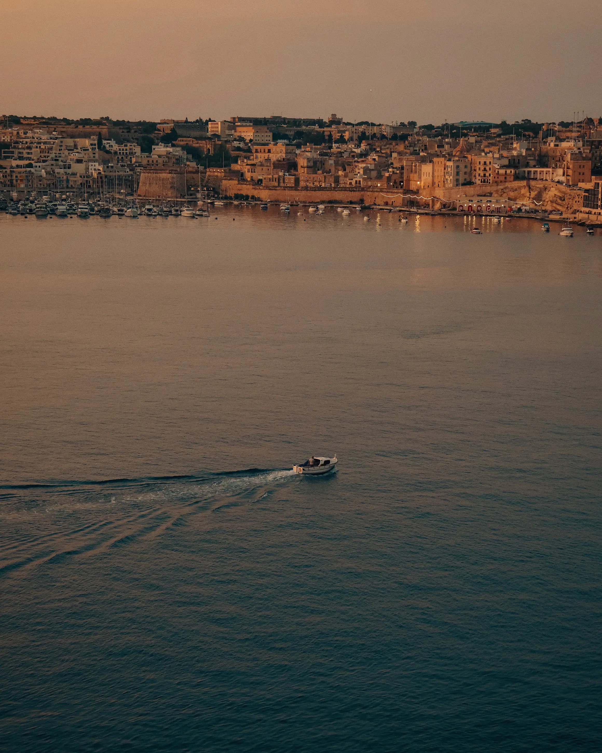 Small boat crossing a calm harbour at sunset in Malta, with historic limestone buildings and a marina illuminated by golden evening light in the background.