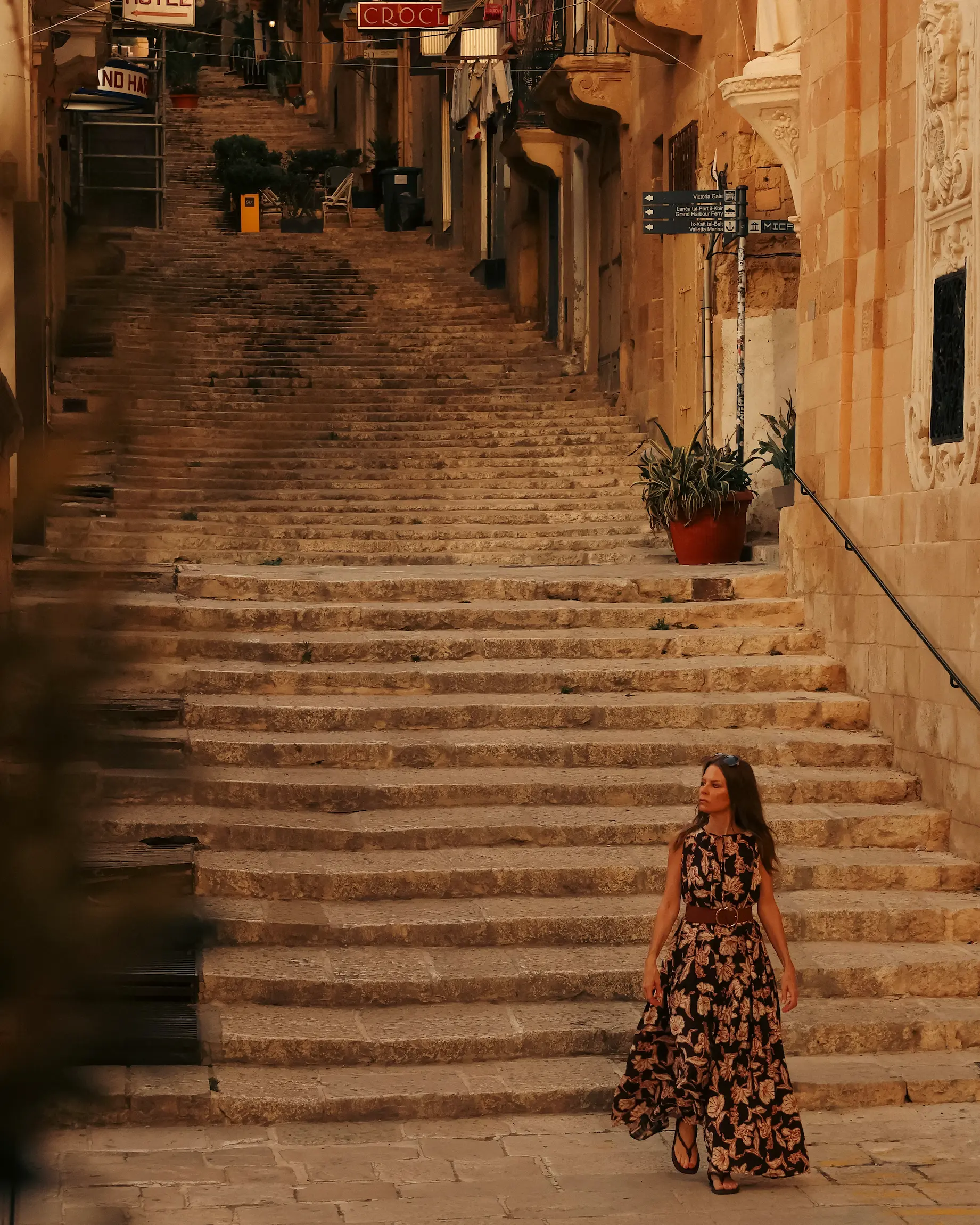 A woman walking through a historic stone square in Valletta, Malta, surrounded by honey-coloured buildings and steep limestone steps illuminated by golden evening light.