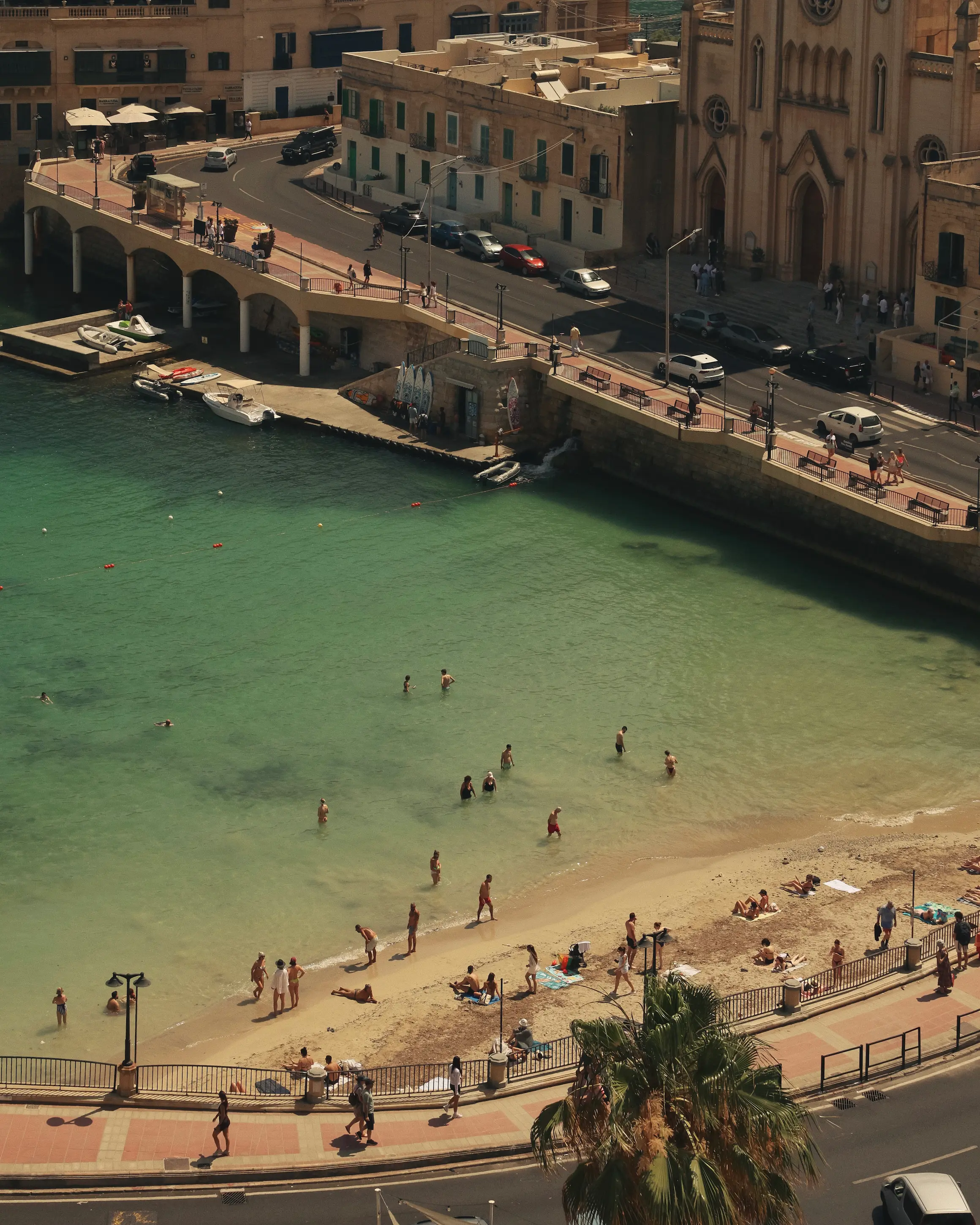 St Julian's beach in Malta with swimmers in turquoise water, a waterfront promenade and historic limestone buildings overlooking the bay.