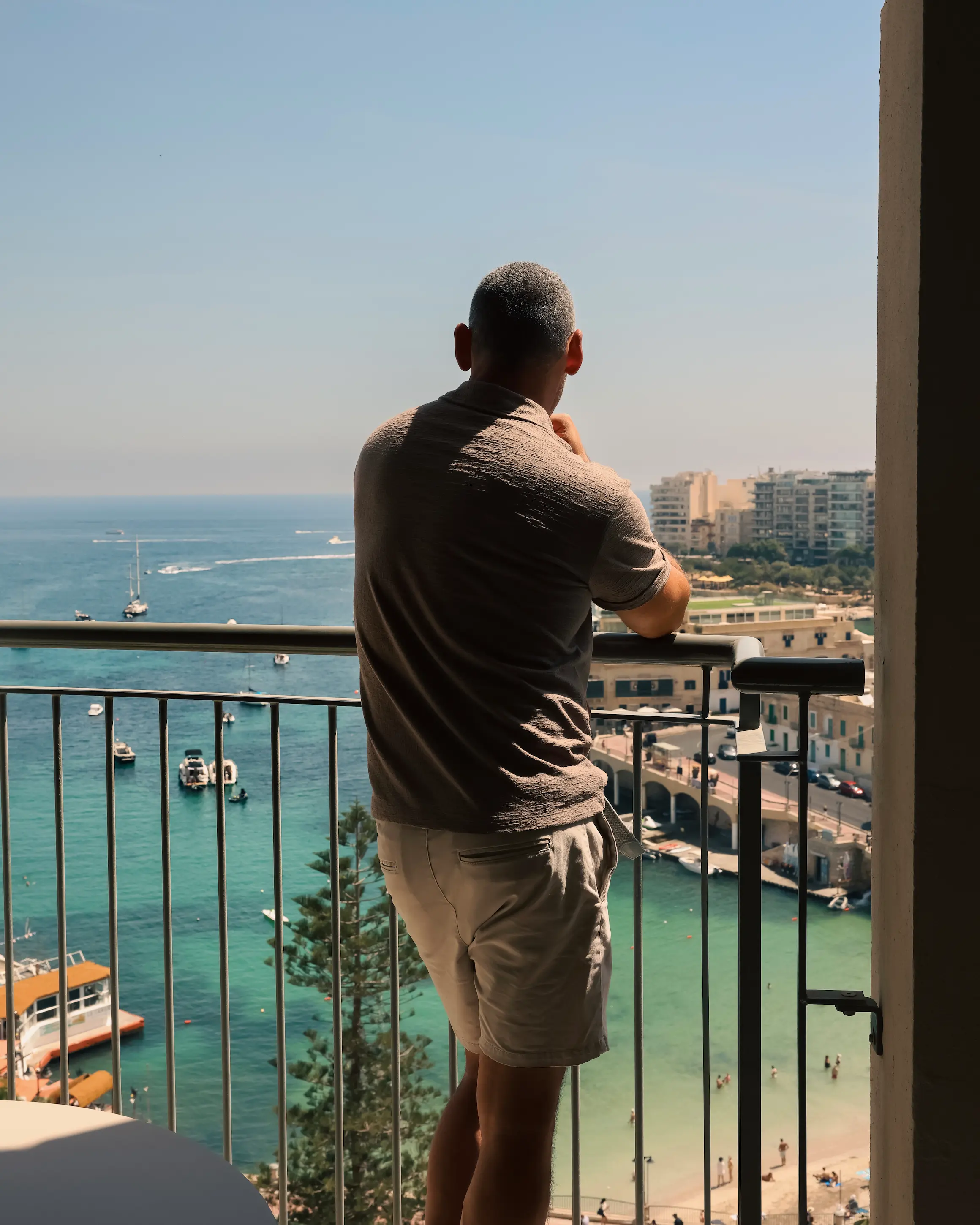 A traveller standing on a hotel balcony overlooking turquoise water in St Julian's Bay Malta, with boats anchored and limestone buildings lining the waterfront below