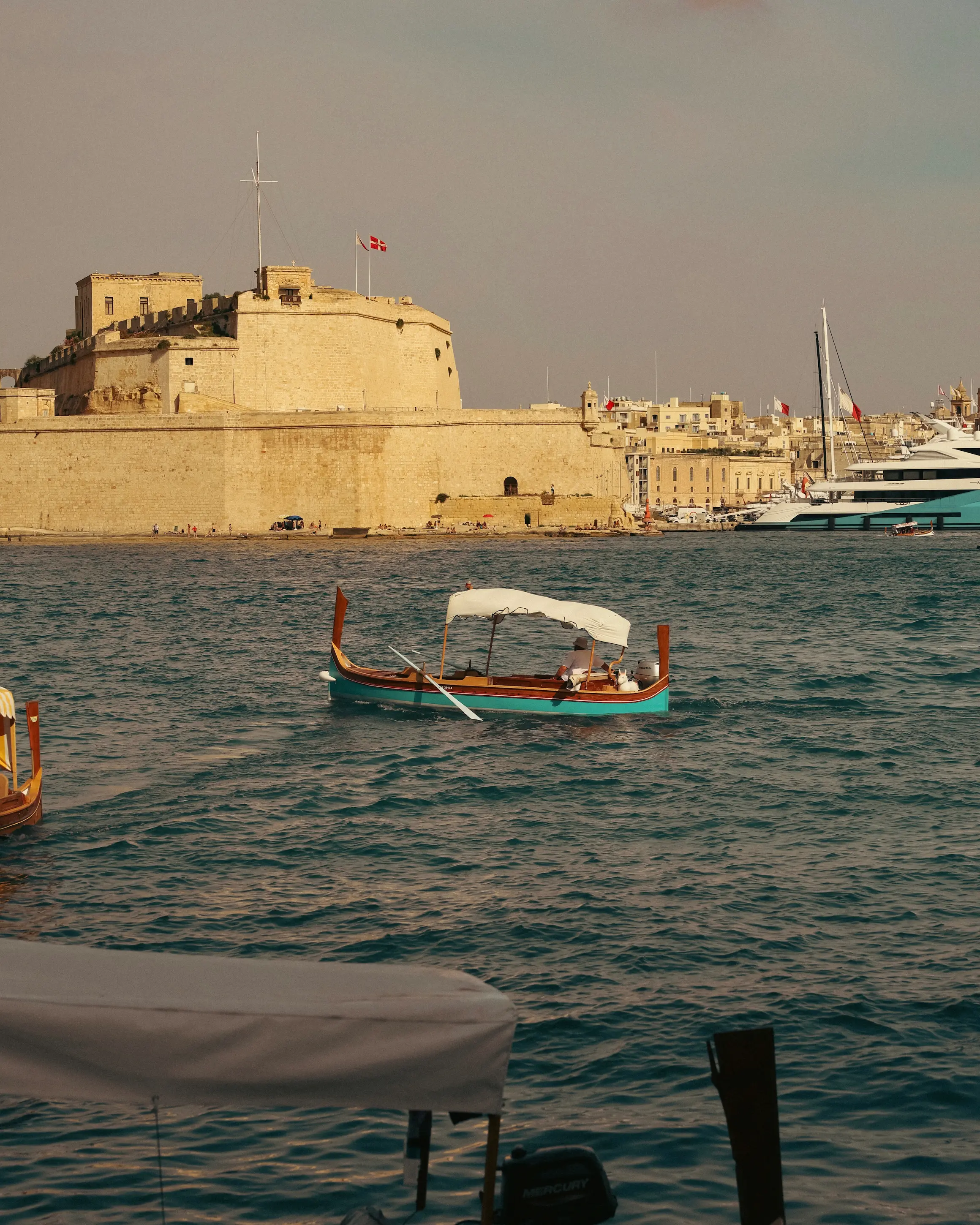 Traditional Maltese boat crossing the Grand Harbour in Valletta, Malta, with historic limestone fortifications and waterfront buildings in the background.