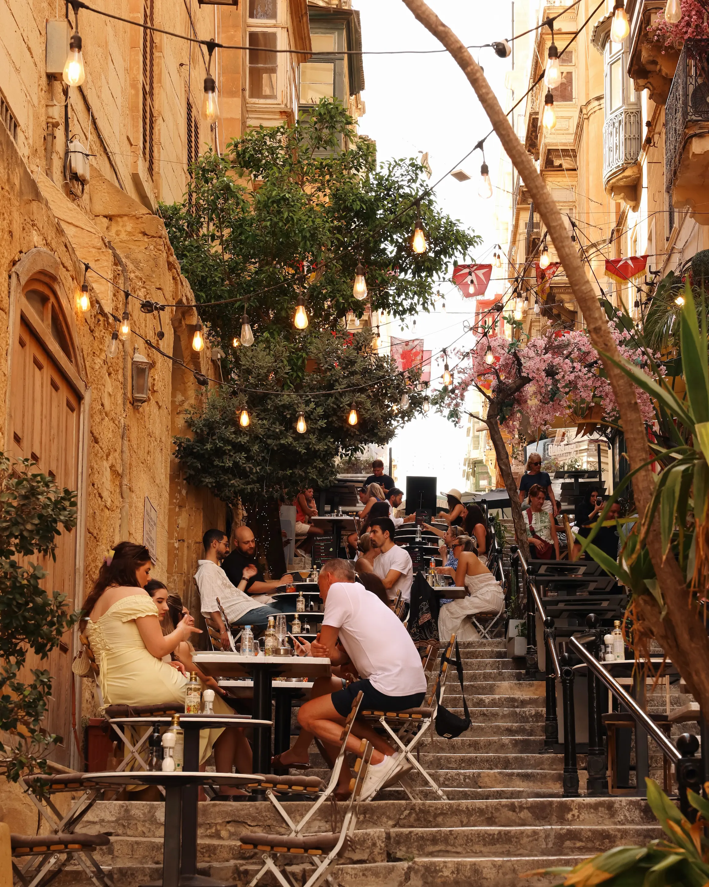 Outdoor diners sit along the famous steps of St Lucia Street in Valletta, Malta, beneath string lights and historic honey-coloured buildings during golden hour.