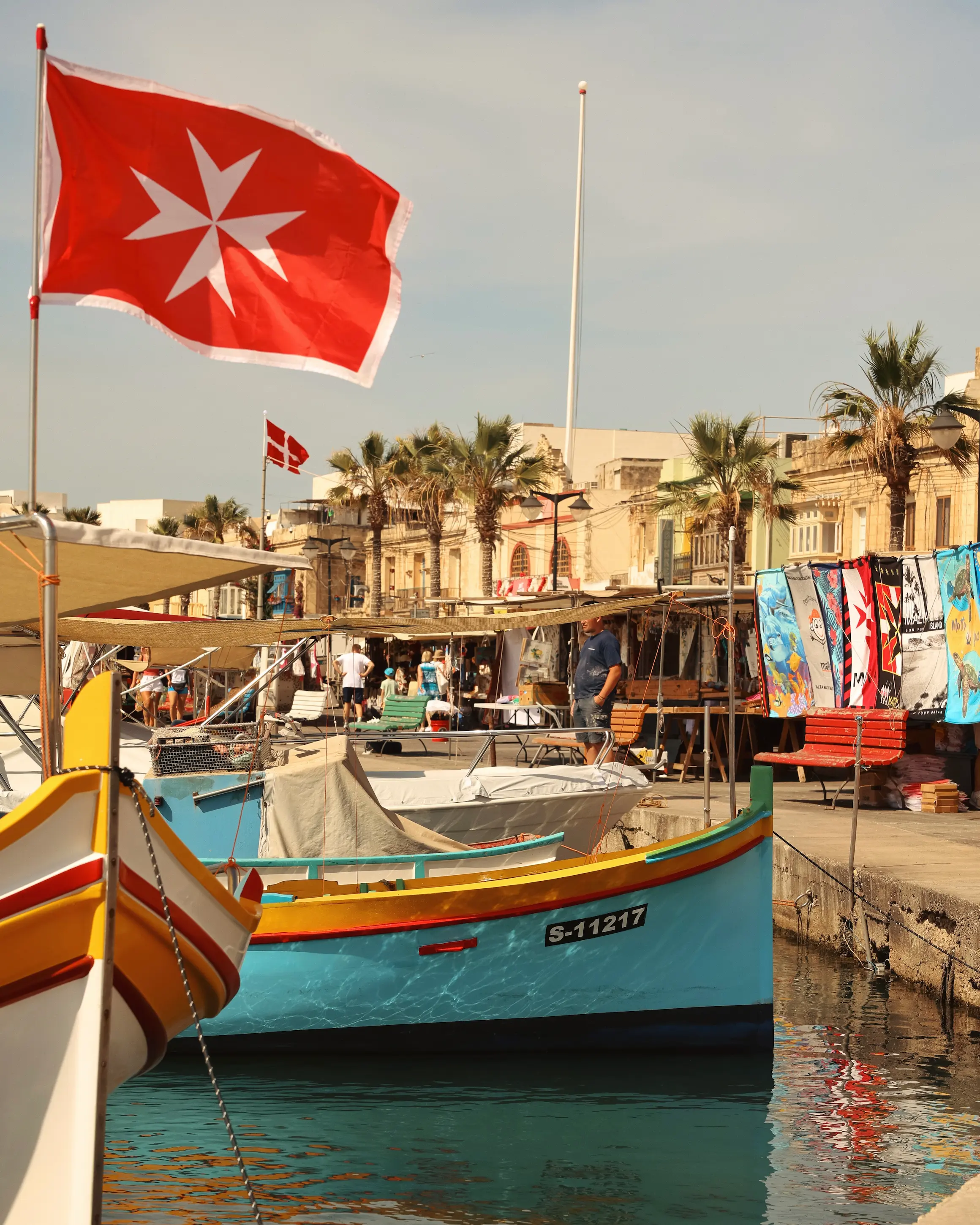 Traditional colourful Maltese fishing boats moored in Marsaxlokk harbour beneath the Maltese flag on a sunny day.
