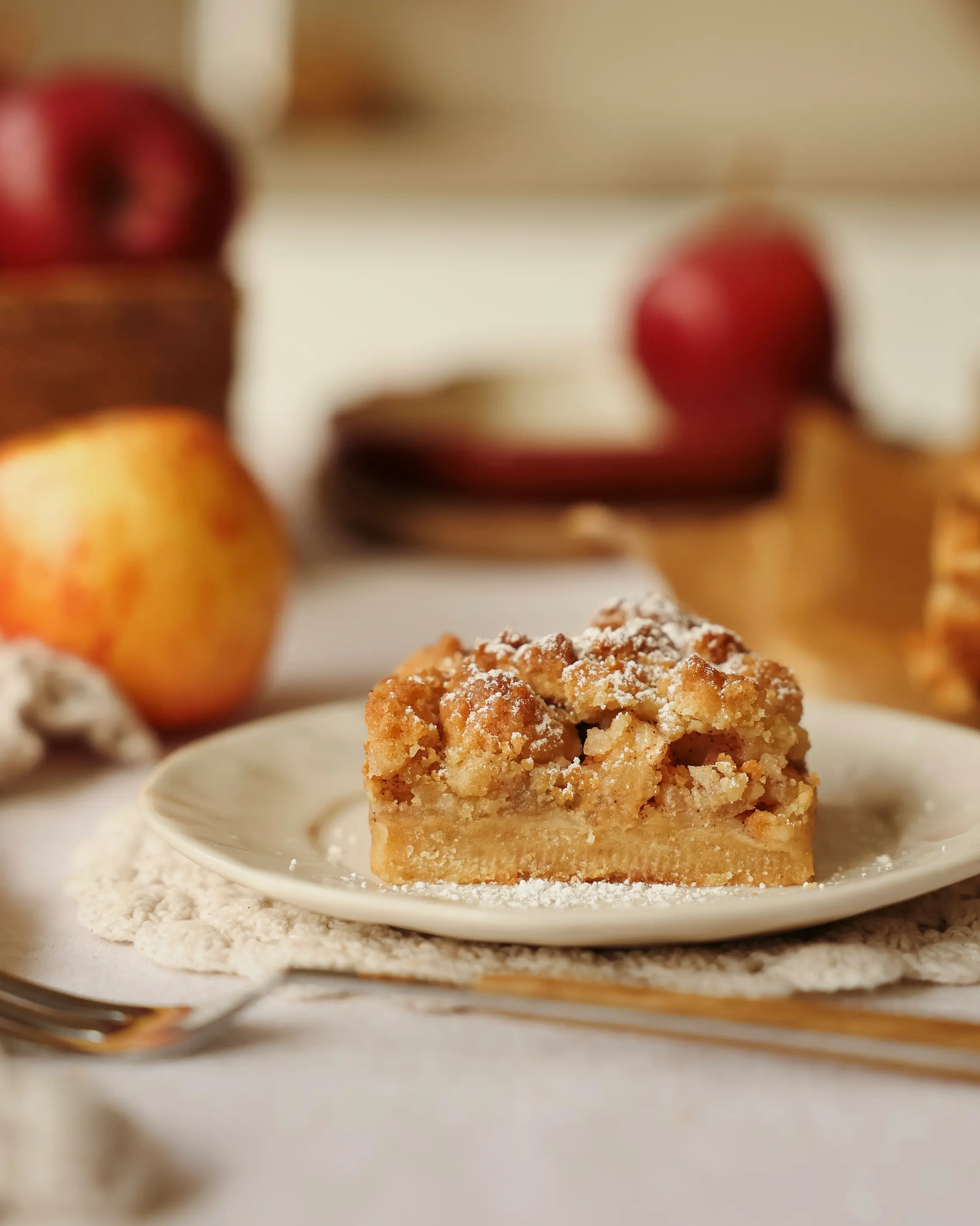 Close-up of a homemade apple crumble bar with a buttery shortbread-style base, cinnamon apple filling and golden crumble topping dusted with icing sugar, served on a cream plate with red apples in the background.