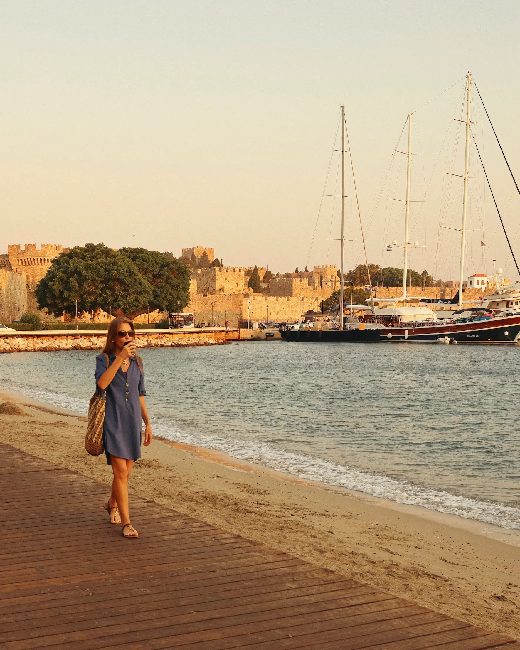 Woman walking along the waterfront in Rhodes Town, with the harbour, yachts and medieval city walls in the background.