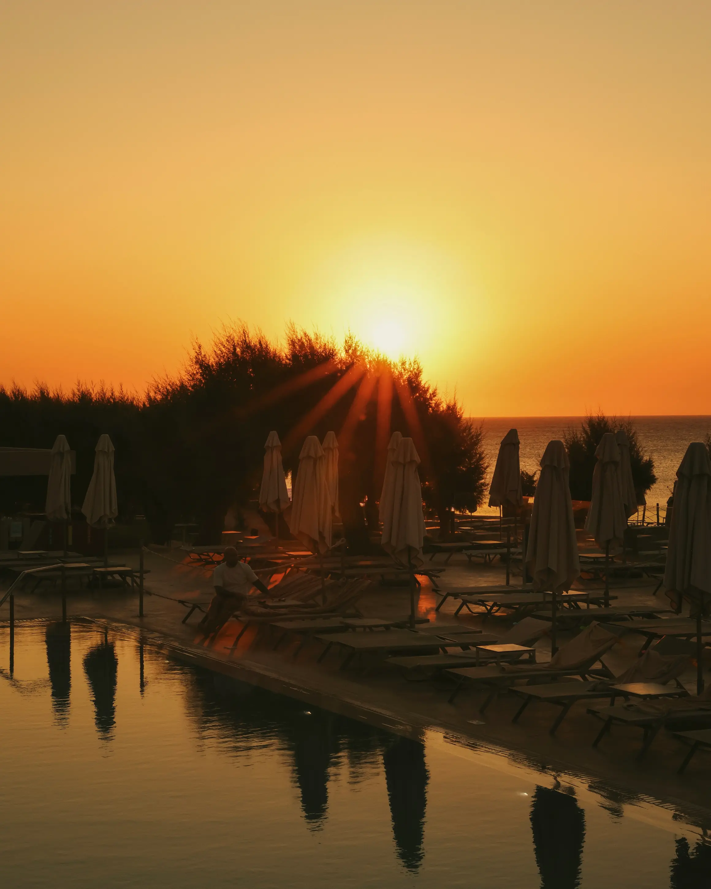 Sunset over the swimming pool and Aegean Sea at Amada Colossos Resort in Rhodes, with sun loungers reflected in the water.