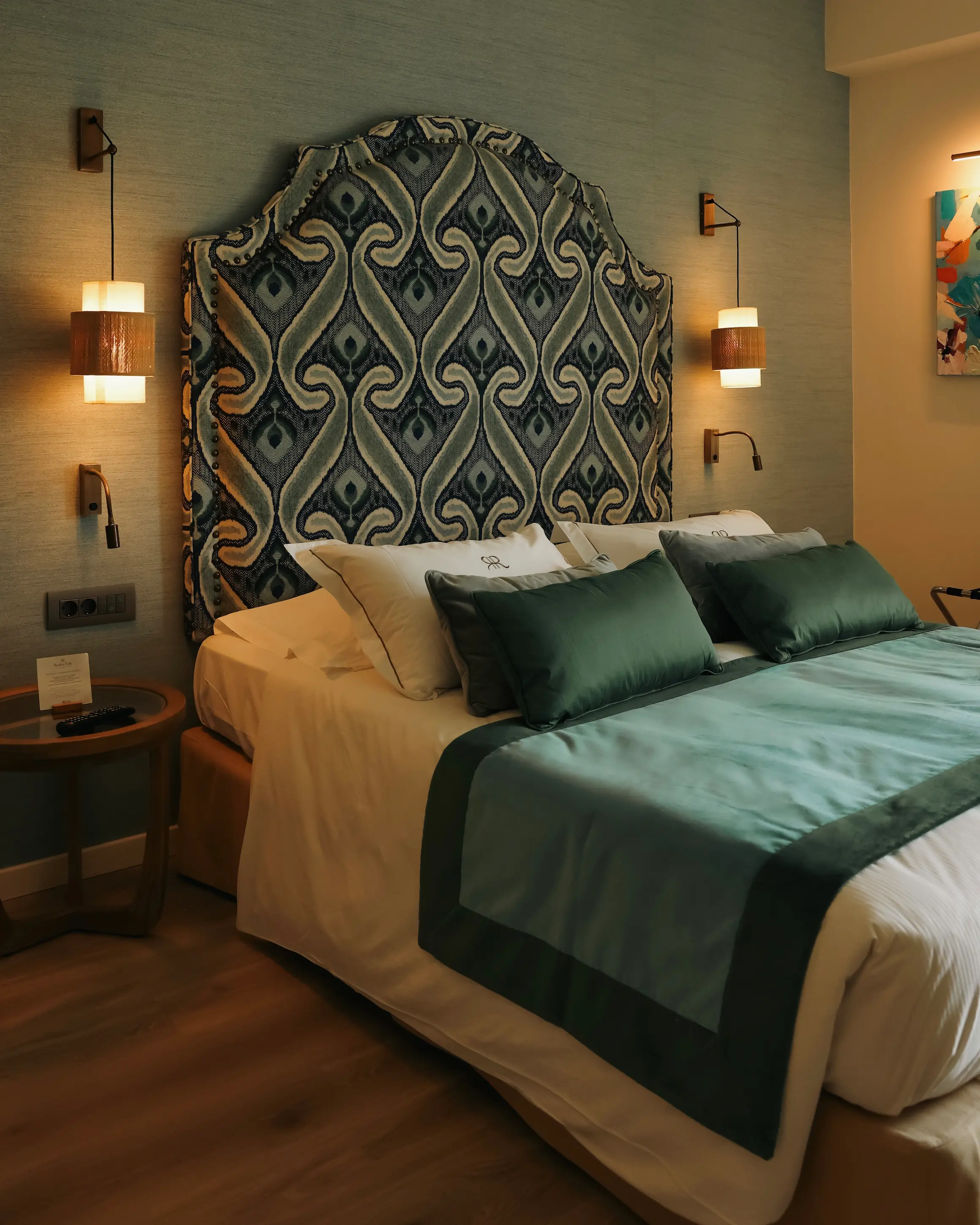 Boutique guest room at Rodos Park in Rhodes, with a large patterned upholstered headboard, teal bedding and warm bedside lighting.