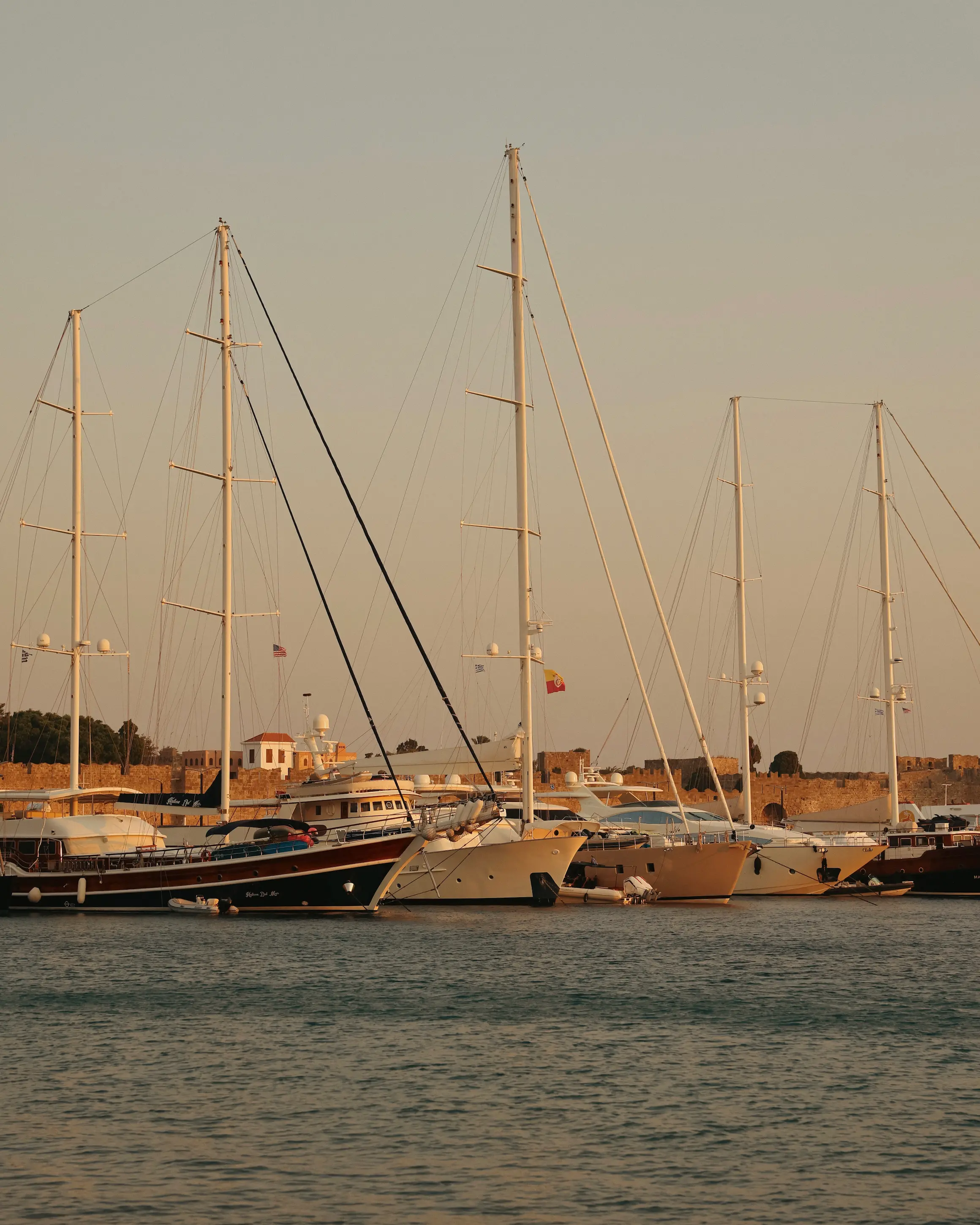 Yachts moored in Mandraki Harbour in Rhodes Town at golden hour, with the medieval city walls in the background.