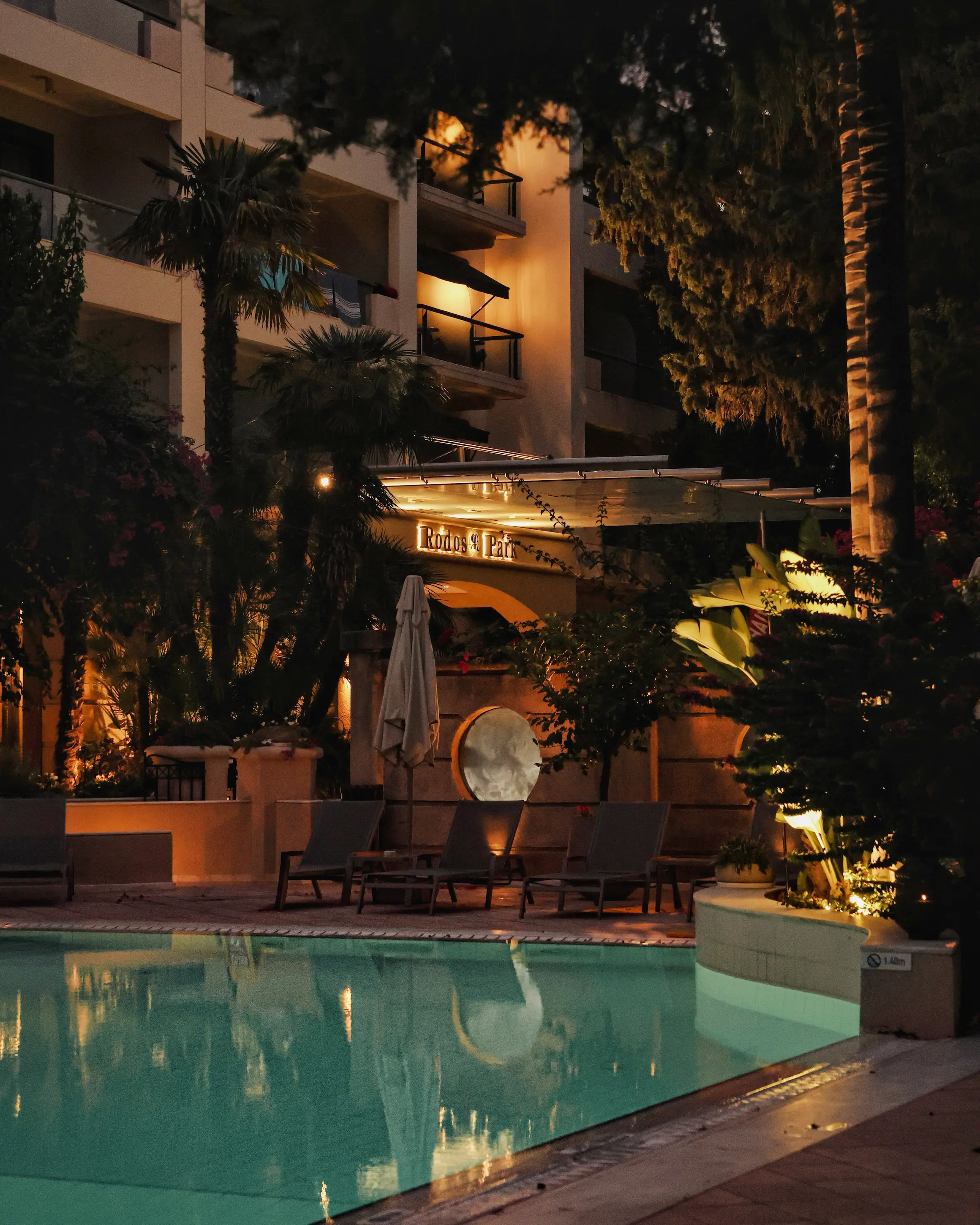Rodos Park hotel pool in Rhodes at night, surrounded by palm trees, greenery and warm evening lights.