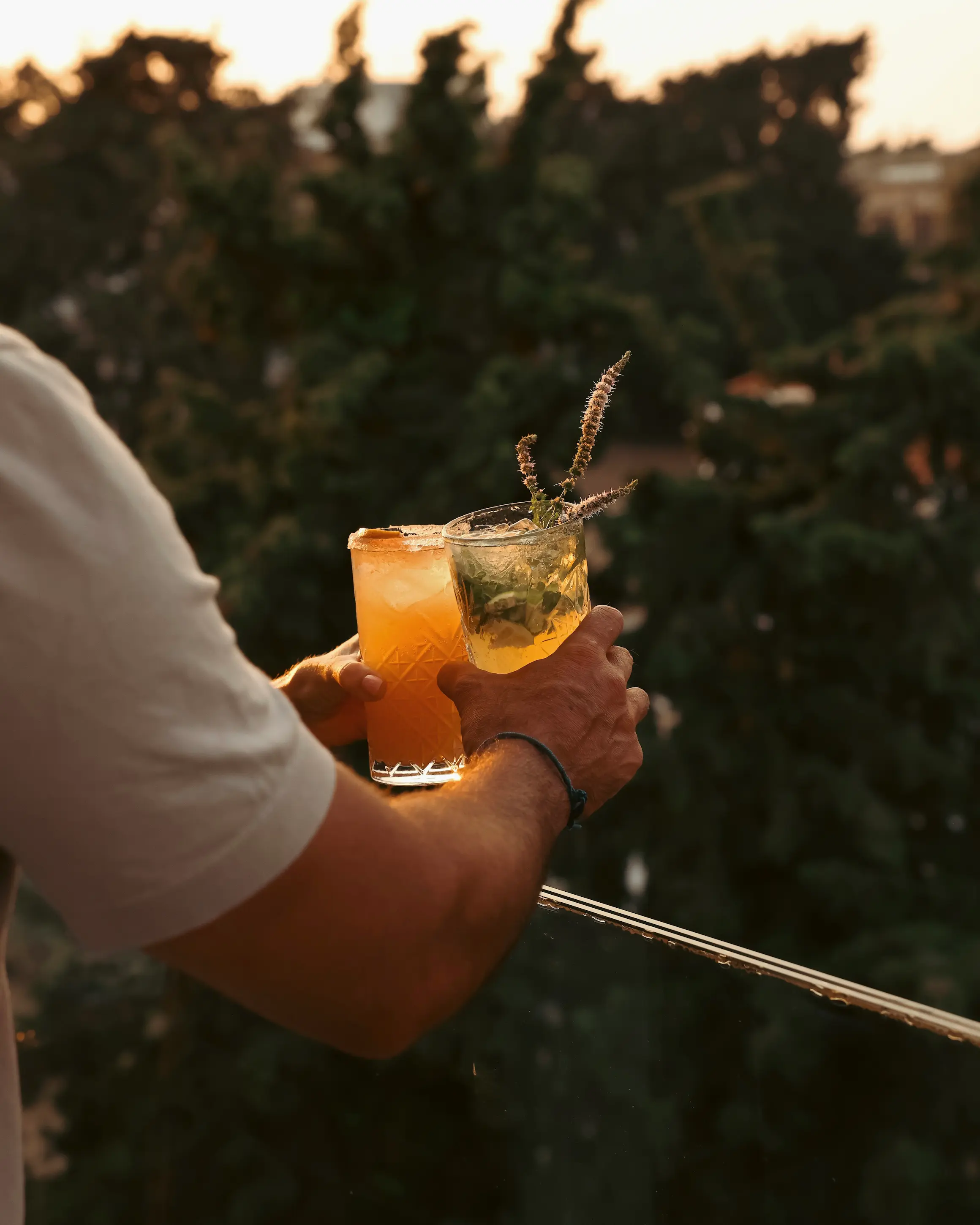 Two cocktails being held at golden hour on the rooftop bar at Rodos Park in Rhodes, with trees and the evening light in the background.