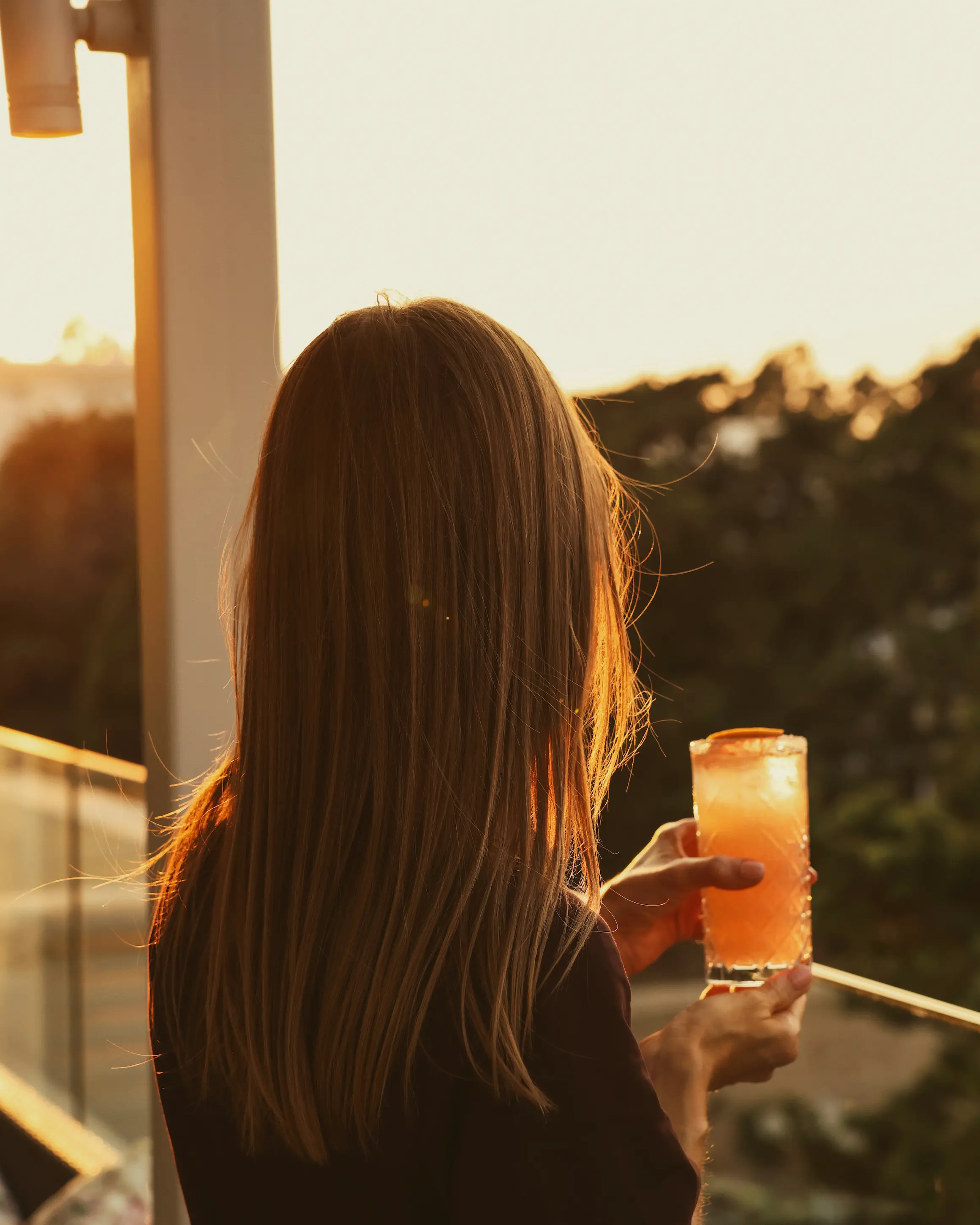 Woman holding a cocktail at golden hour on the rooftop bar at Rodos Park in Rhodes, with warm sunset light over the surrounding trees.