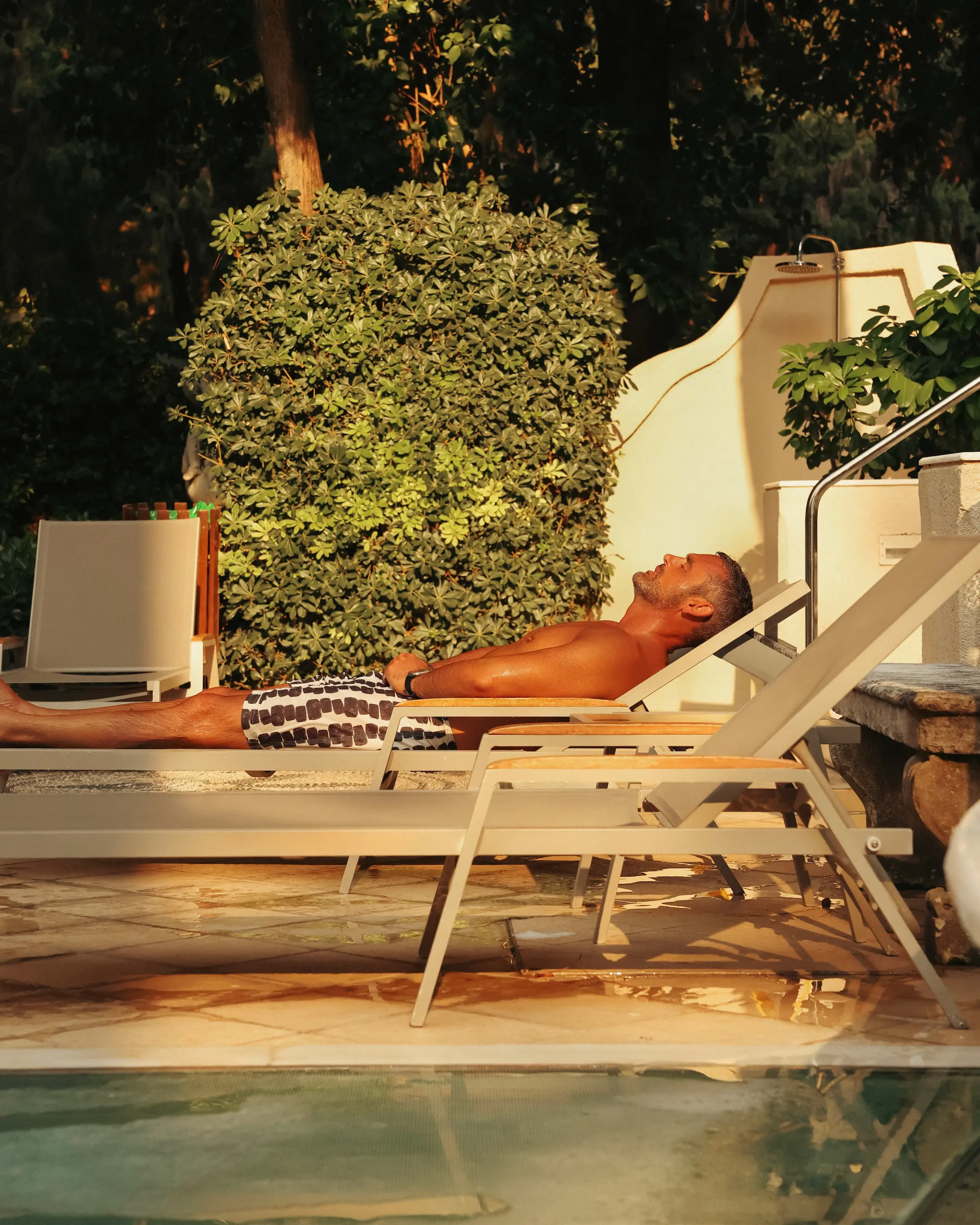 Guest relaxing on a sun lounger beside the pool at Rodos Park in Rhodes, surrounded by greenery in the late afternoon sun.