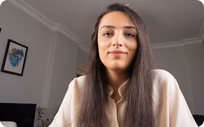 Young woman with long straight dark hair, wearing a light-colored blouse, looking directly at the camera in a softly lit indoor setting with framed artwork and a plant in the background.