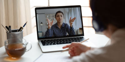 Person participating in a video call on a laptop, gesturing with both hands while another person listens with headphones.