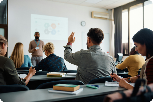 A classroom with an instructor standing near a screen presenting a blurred chart, and students seated facing the front; one student raises his hand while others take notes or listen.