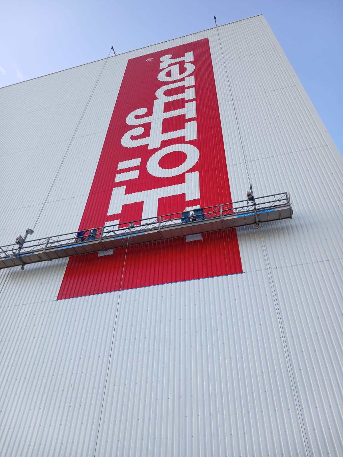 Monteure bringen den roten Höffner-Schriftzug aus der Seilkorbanlage an der Hochregallager-Fassade an