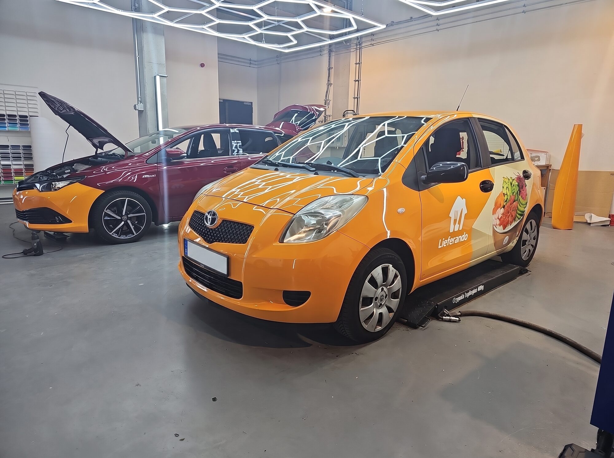 Toyota Yaris in Lieferando-Orange, im Hintergrund ein weiteres Fahrzeug in Arbeit