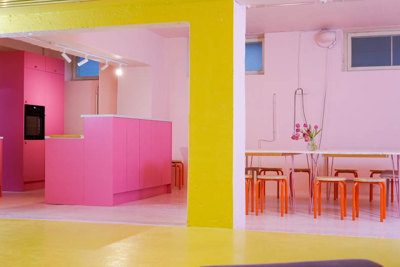 Modern interior with bright pink kitchen cabinets, orange stools, white tables, yellow floor, and pink walls.