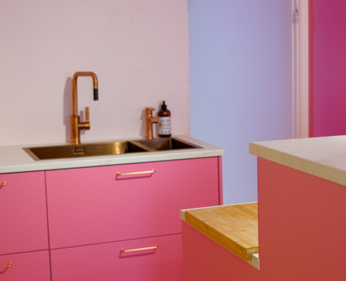 Modern kitchen with pink cabinets, white countertop, copper faucet, and a wooden cutting board.